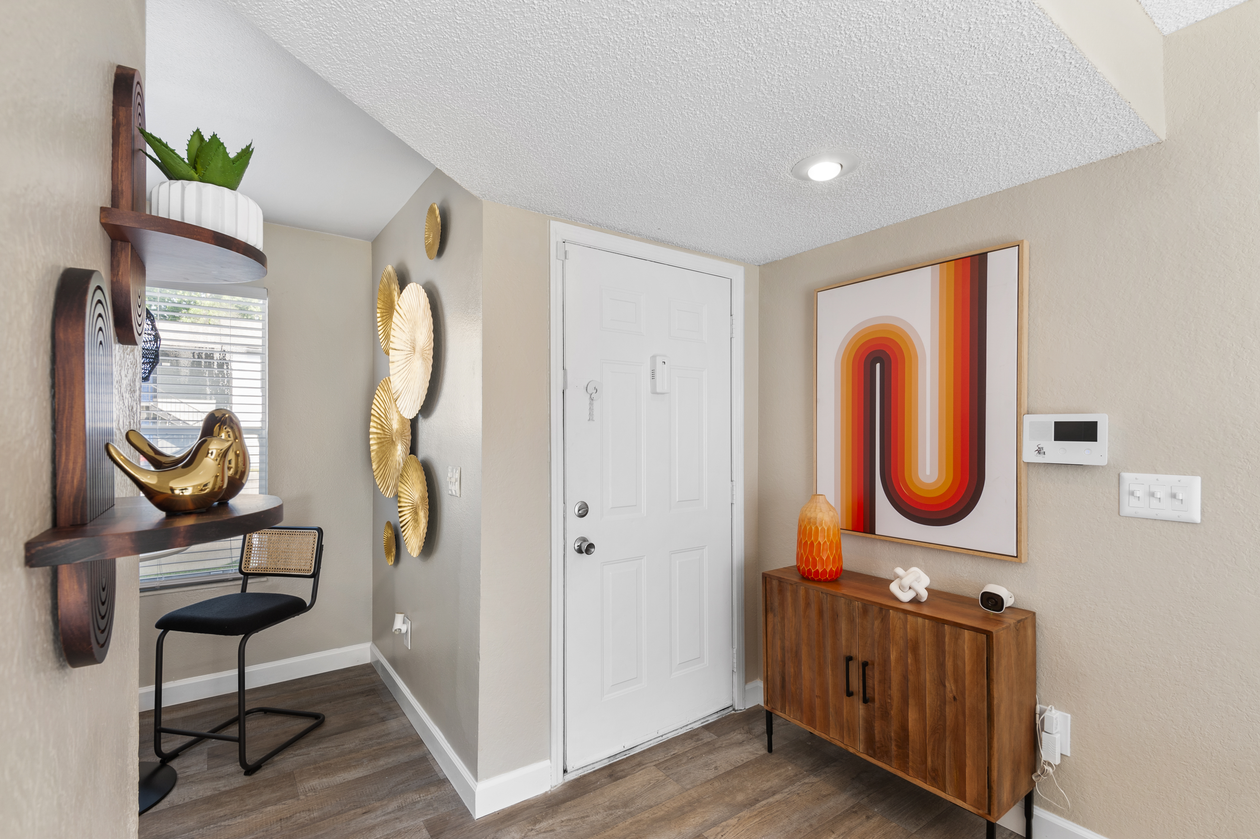 A modern entryway featuring a light-colored wall with decorative art and wall hangings. There's a wooden cabinet with decor pieces, a stylish chair, and a door leading outside. Natural light streams in through a window, highlighting the warm tones of the artwork and furnishings.