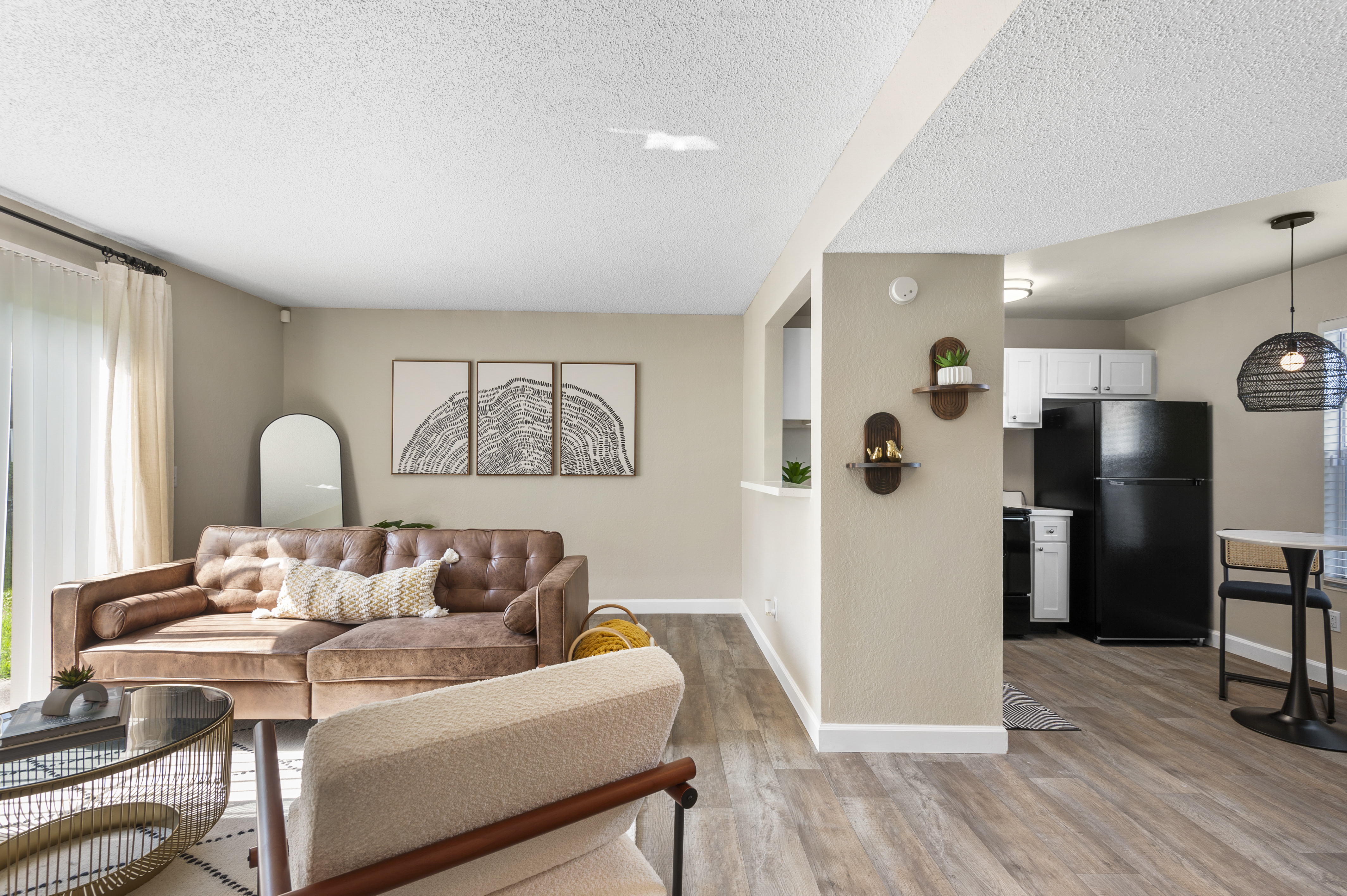 A bright and modern living space featuring a brown leather sofa, a round coffee table, and a large mirror. There are abstract wall art pieces and a small plant on the wall. In the background, a kitchen area is visible with black appliances and white cabinets, creating a cozy and inviting atmosphere.