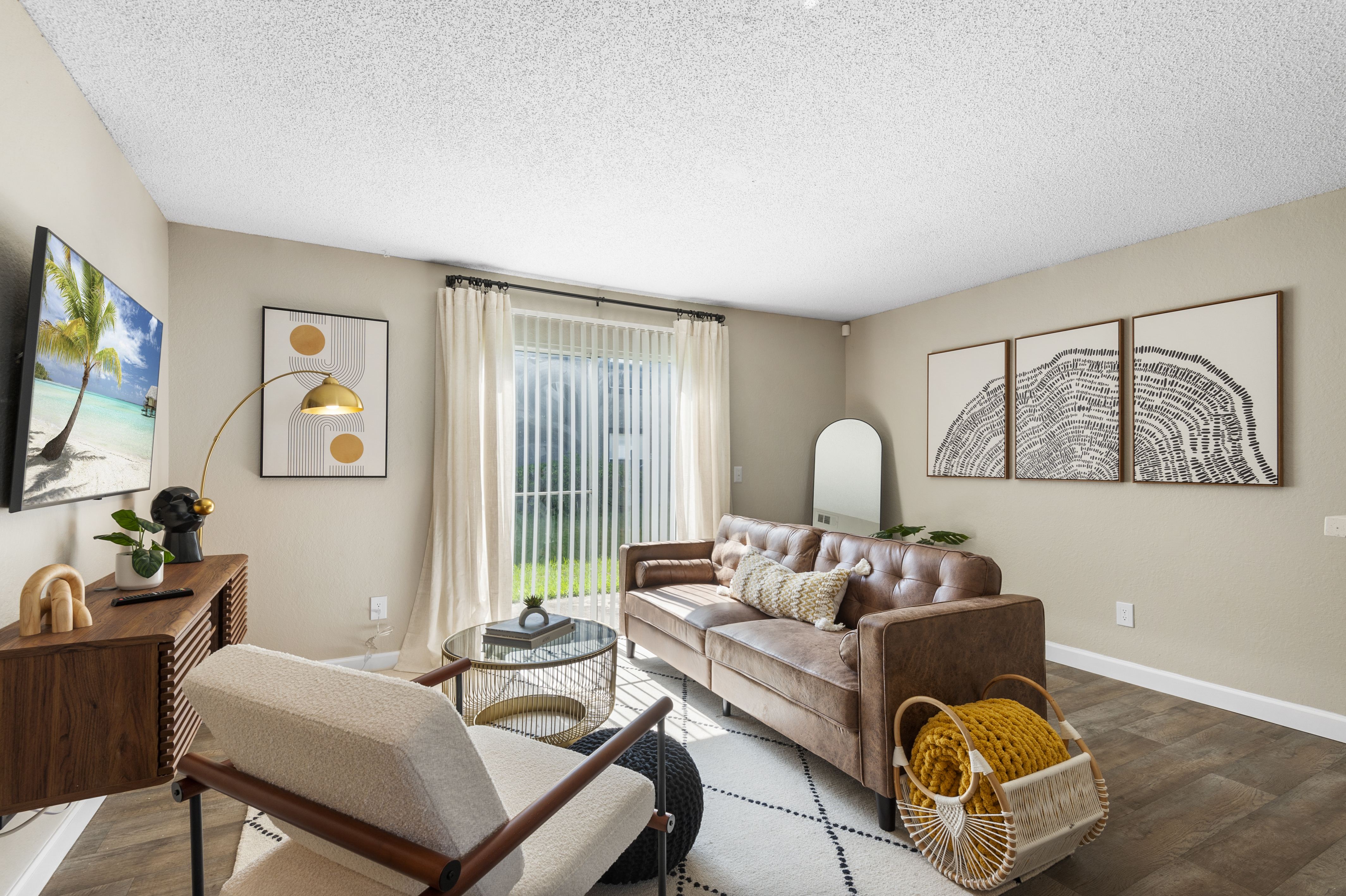 A cozy living room featuring a brown leather sofa, a modern armchair, and a wooden sideboard. Large windows provide natural light, with sheer curtains softening the view. Two framed artworks hang on the wall, and a decorative basket sits beside the armchair, enhancing the room's inviting atmosphere.