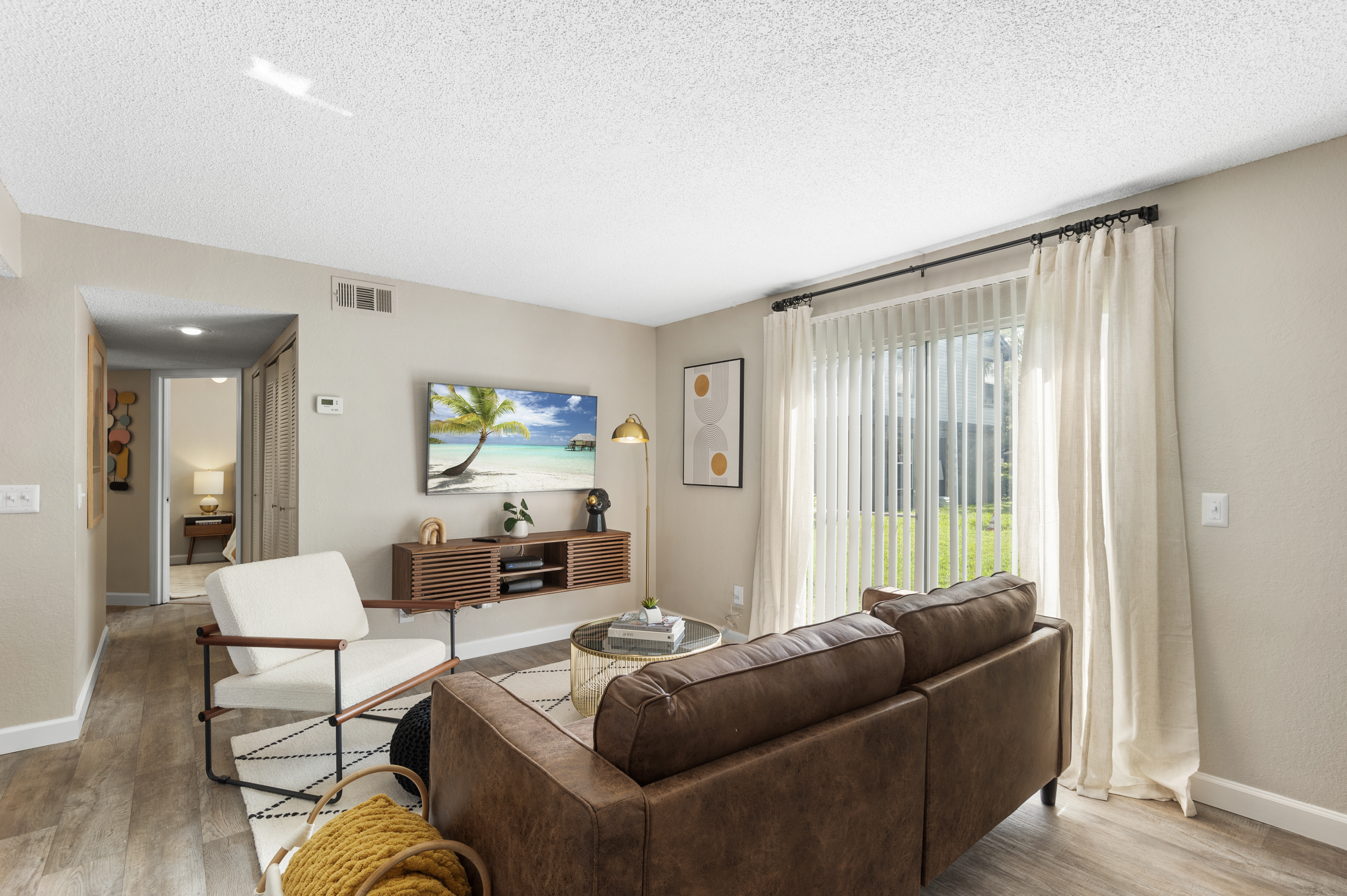 Modern living room featuring a brown leather sofa, a white armchair, and a stylish coffee table. A large TV is mounted on the wall, displaying a beach scene. Natural light filters through sliding glass doors, with sheer curtains. The room has a warm color palette and wooden flooring, creating an inviting atmosphere.