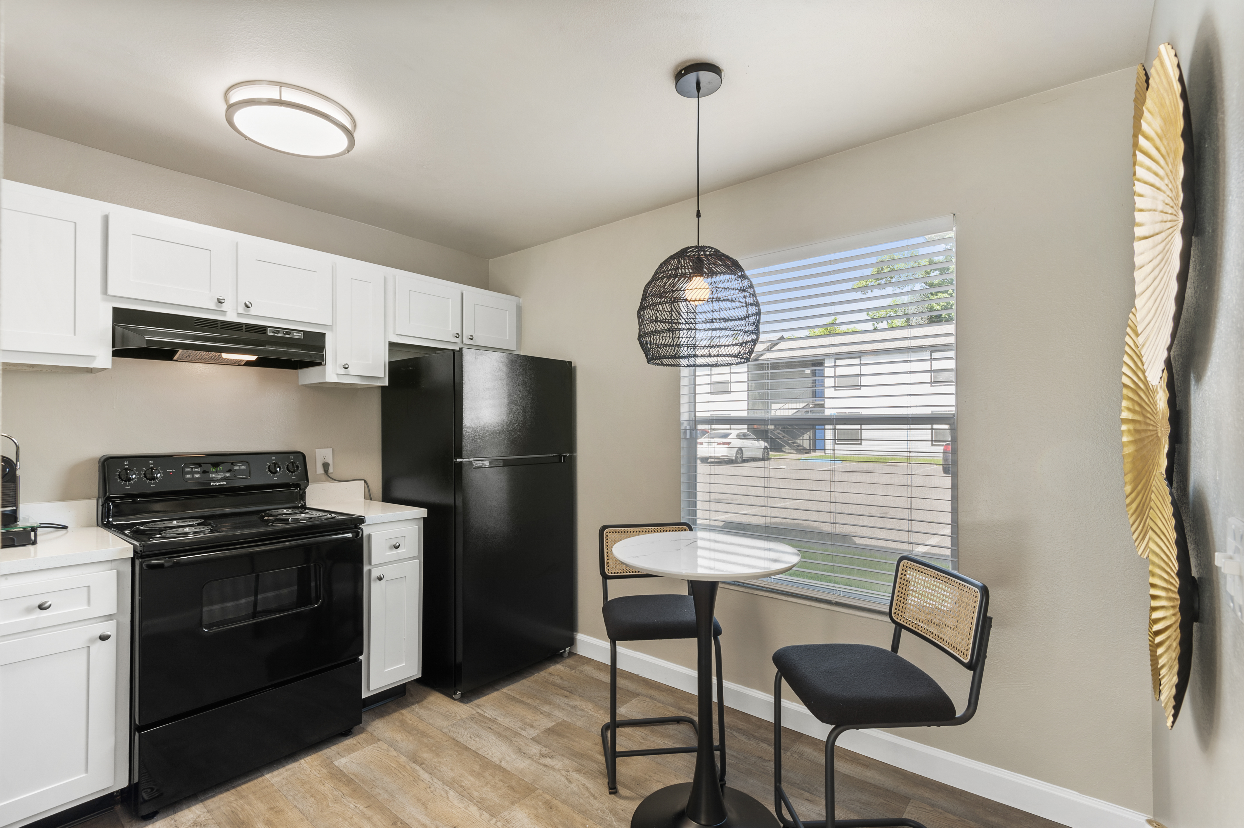 A modern kitchen featuring white cabinetry, a black stove and refrigerator, and a small dining area with a round table and two black chairs. Natural light comes through a window with blinds, and a pendant light hangs above the table. The design is contemporary and functional.