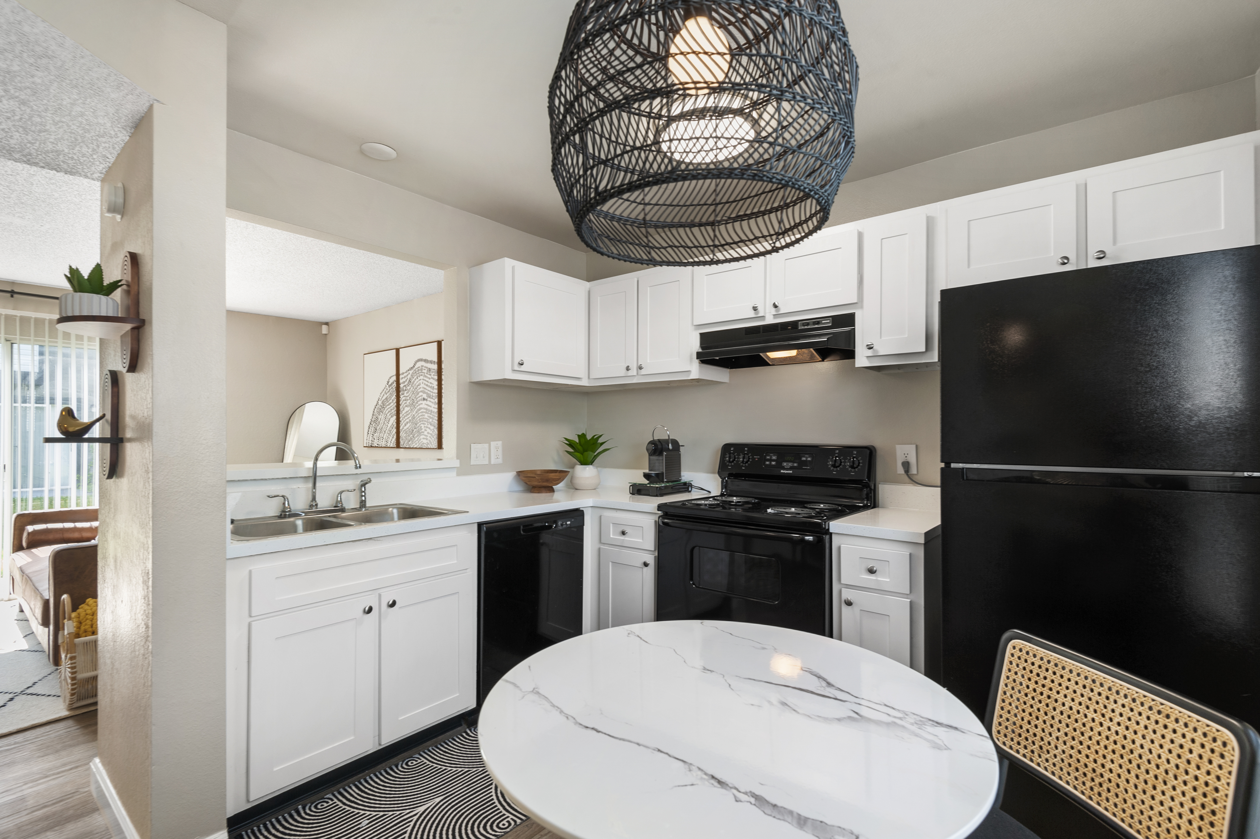 A modern kitchen featuring white cabinetry, a black refrigerator, and a stove. A round marble dining table with a woven chair is positioned under a large pendant light. The space is well-lit with natural light, and there are decorative plants and artwork adding to the cozy atmosphere.