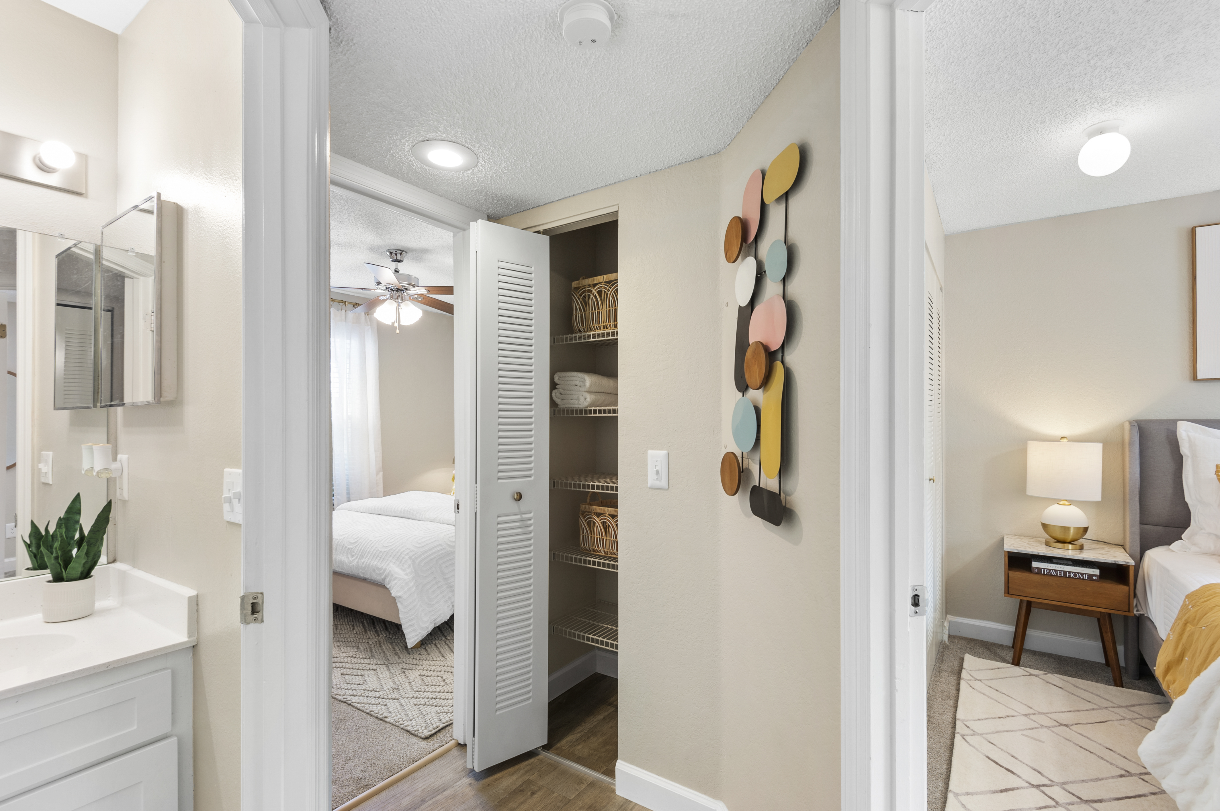 Interior view of a modern bedroom and bathroom entrance. The bathroom features a vanity with a mirror and a potted plant, while the adjacent room has a bed, nightstand, and decorative wall art. A closet is partially visible, lined with baskets. Soft lighting and neutral colors create a warm atmosphere.