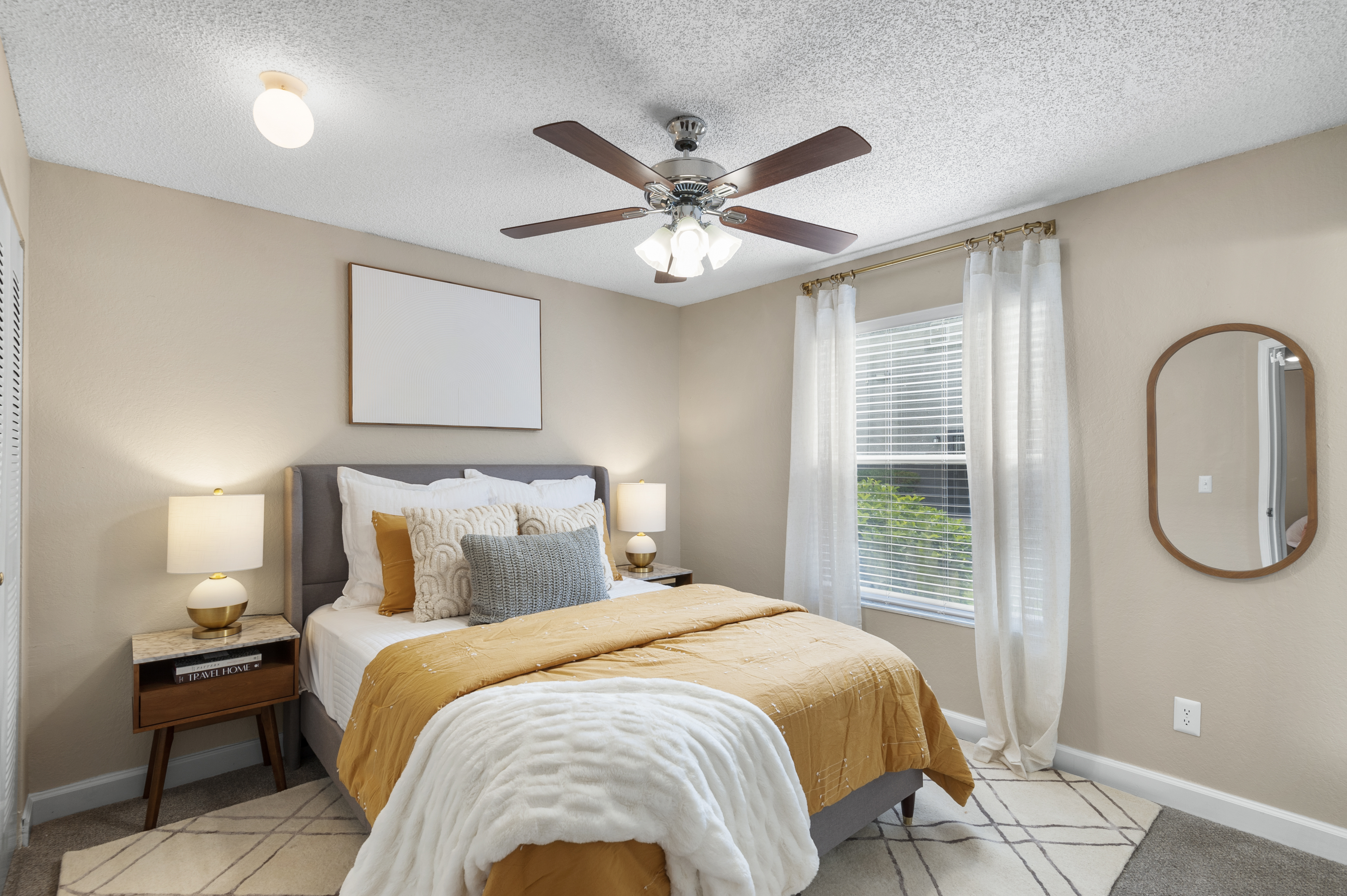 A cozy bedroom featuring a queen-sized bed with yellow and white bedding, a modern ceiling fan, two bedside lamps, and a brown nightstand. The walls are a soft beige, and there is a large mirror and a decorative wall art piece. Natural light filters in through the window, adding warmth to the space.