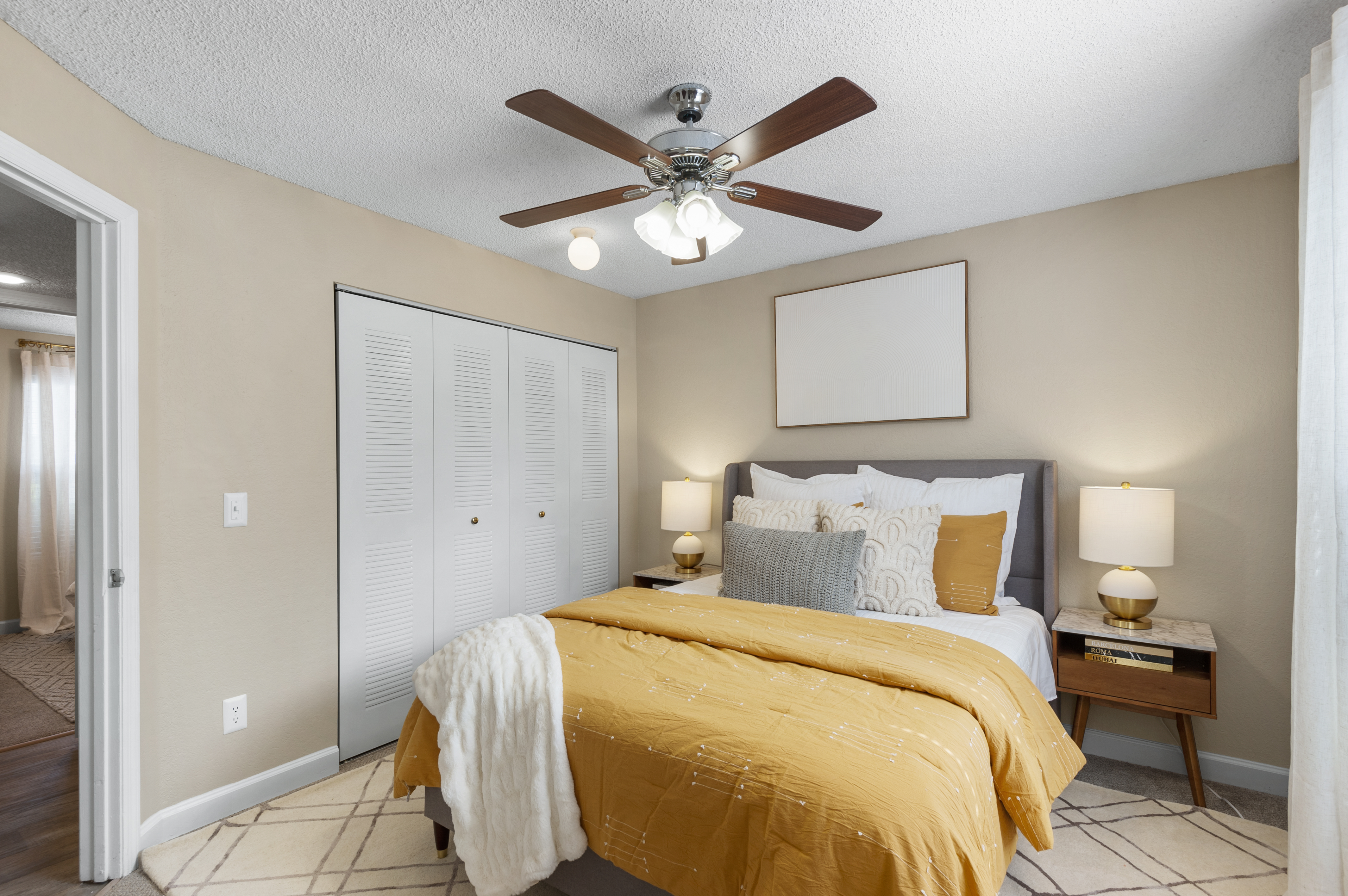 A cozy bedroom featuring a queen-sized bed with yellow and white bedding, two bedside lamps, a stylish wooden nightstand with decor, and a ceiling fan. The walls are painted a soft beige, and there is a large closet with white sliding doors. Natural light filters in through sheer curtains.