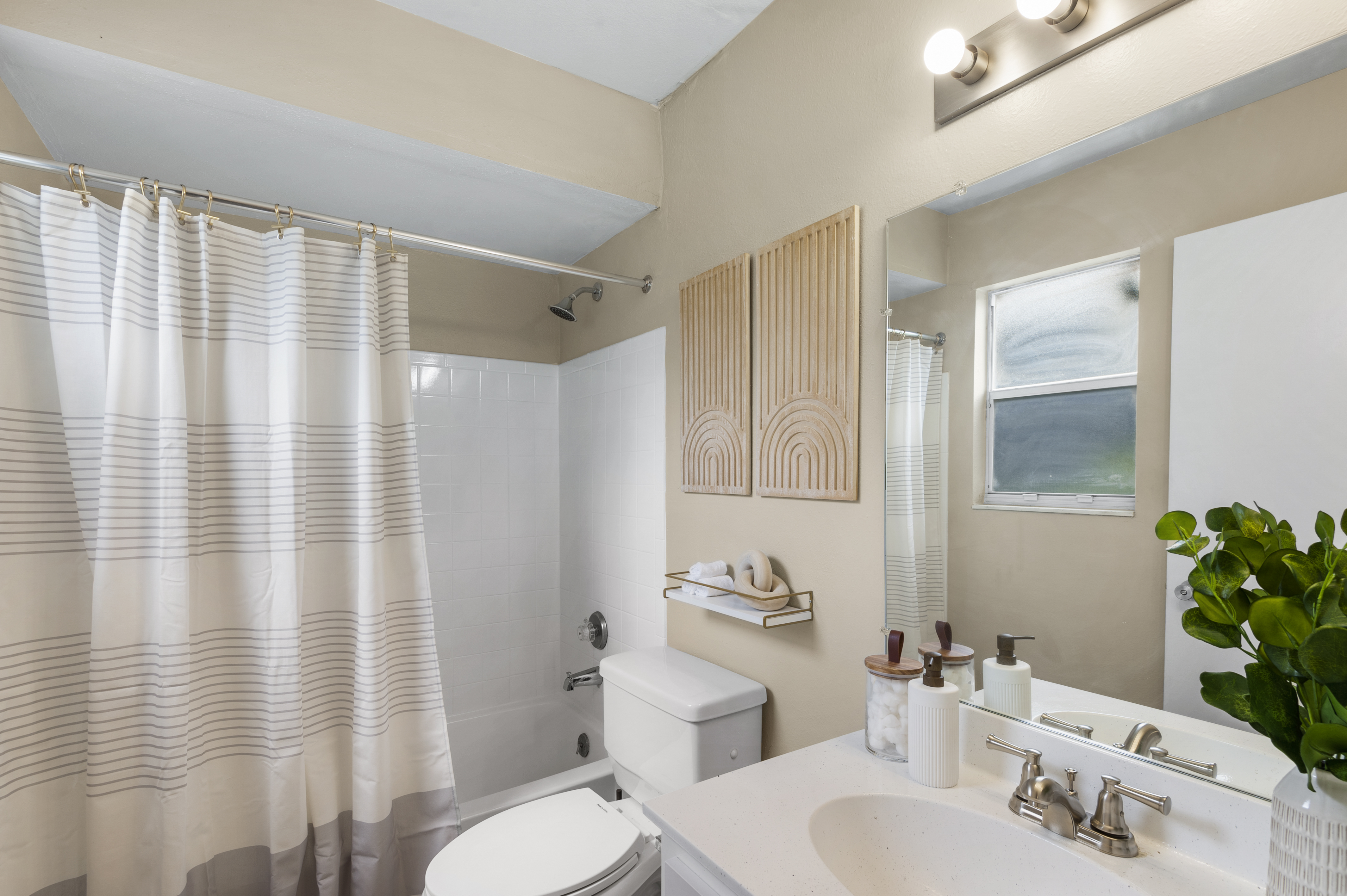 A modern bathroom featuring a white tub with a shower curtain that has horizontal stripes, a sleek toilet, and a minimalist vanity with two sinks. Above the sinks, there are decorative elements, and a small green plant adds a touch of greenery to the space. Natural light shines through a window.