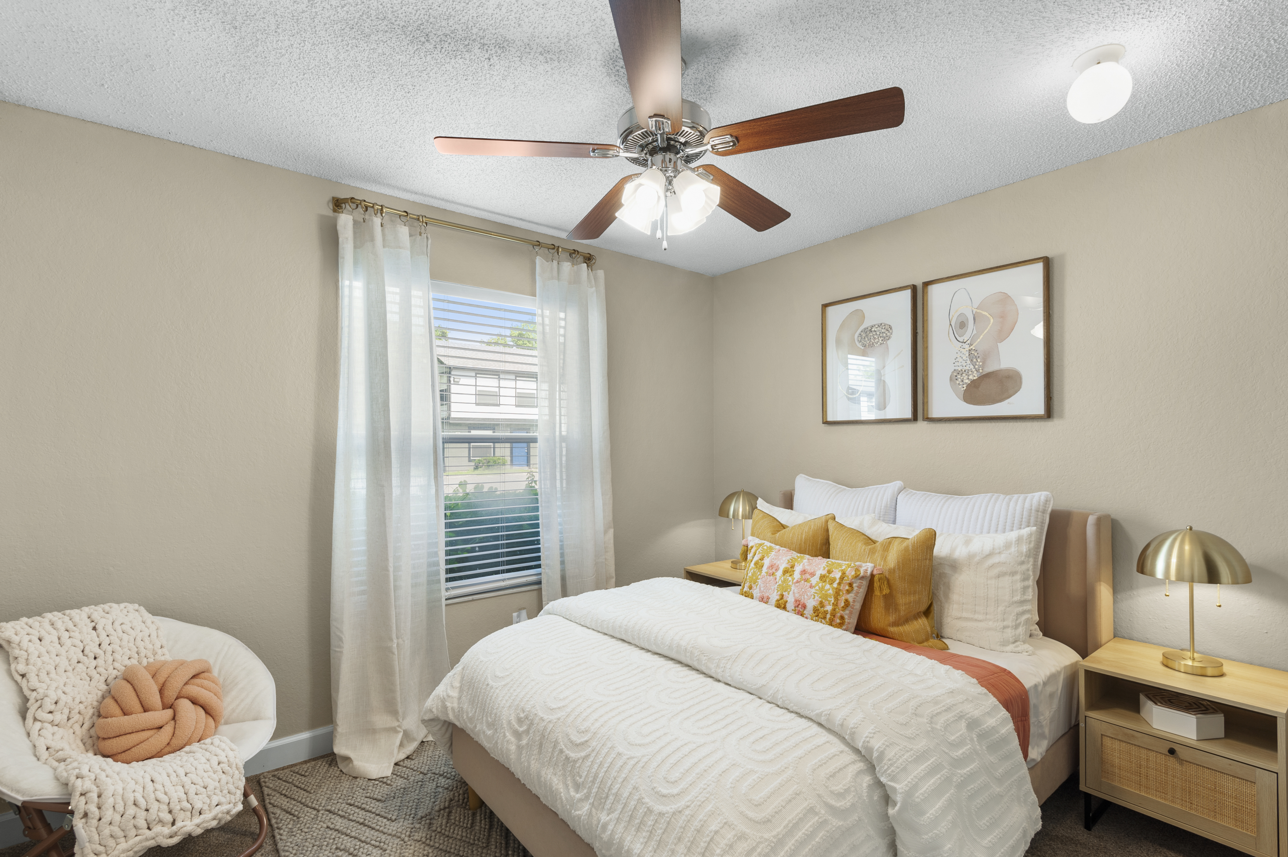 Cozy bedroom featuring a queen-sized bed with white bedding and colorful throw pillows. A stylish ceiling fan with light hangs above. Natural light from a window illuminates the room, complemented by sheer curtains. There's a small nightstand with a lamp and decorative art on the wall, creating a warm atmosphere.