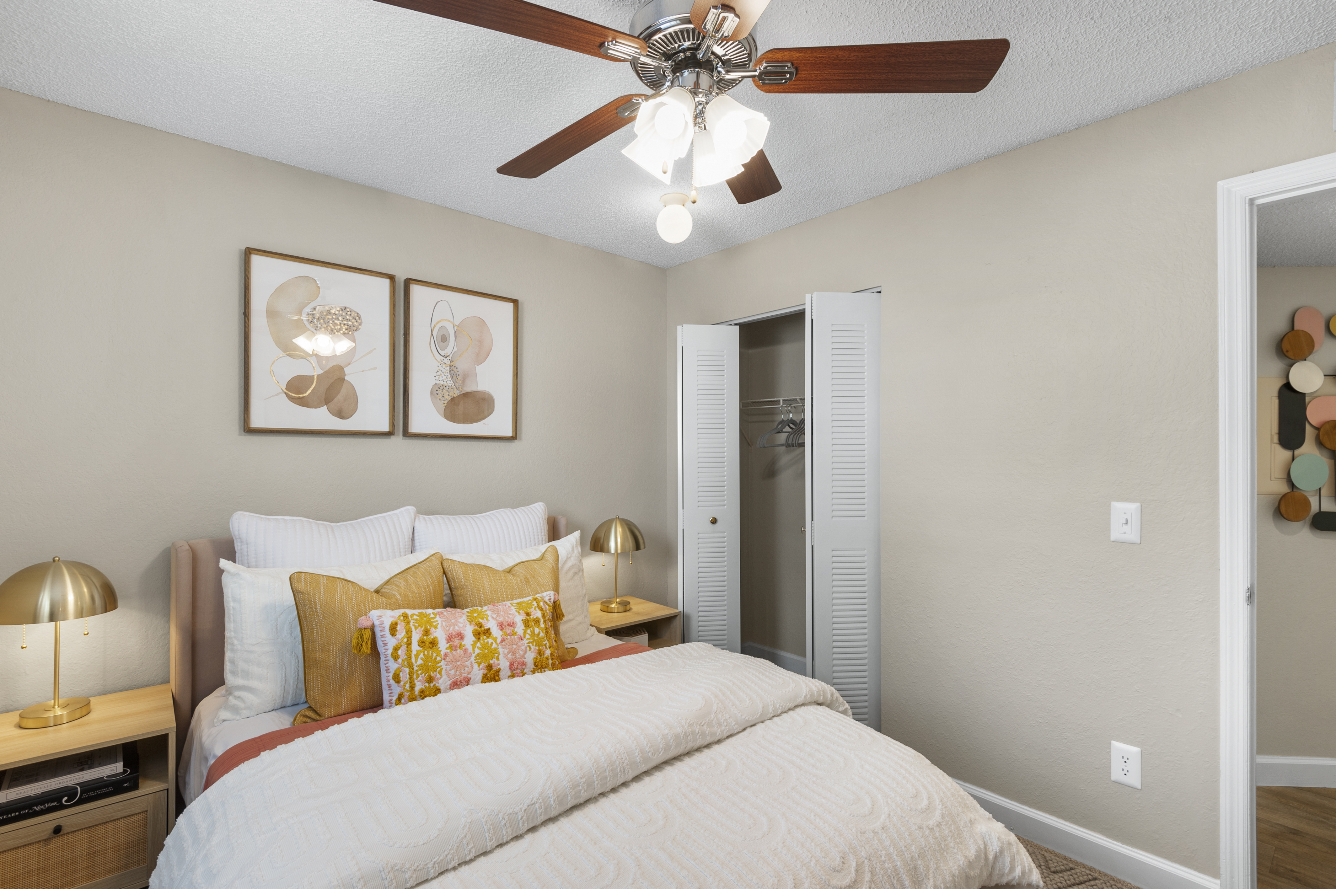 A cozy bedroom featuring a light-colored comforter, decorative pillows, and bedside lamps. A ceiling fan with light fixtures hangs above. The room has a neutral-toned wall, framed artwork, and a closet with sliding doors. Soft furnishings create a warm and inviting atmosphere.