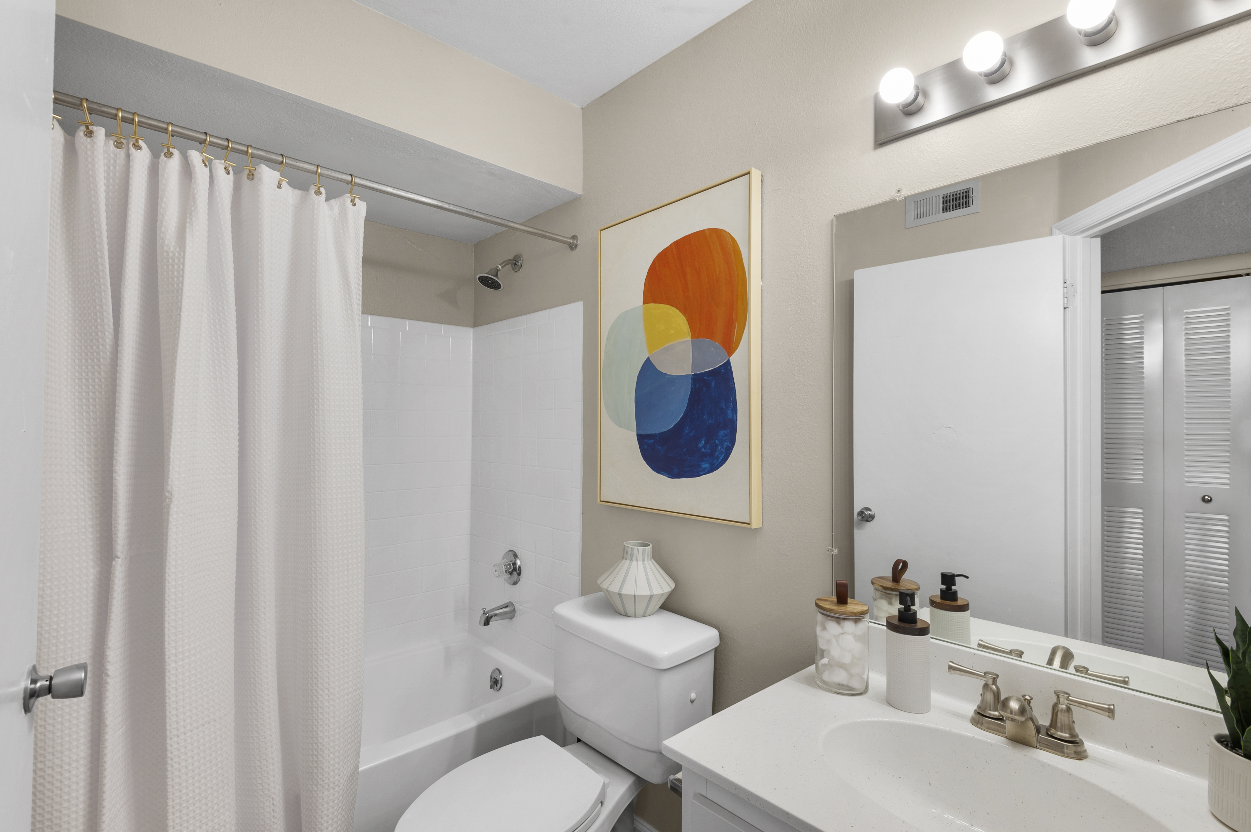 A modern bathroom featuring a white bathtub with a shower curtain, a white toilet, and a vanity with a sink. The wall above the sink is decorated with a colorful abstract painting, and a light fixture with three bulbs is mounted above the mirror. The decor is minimalistic and clean.