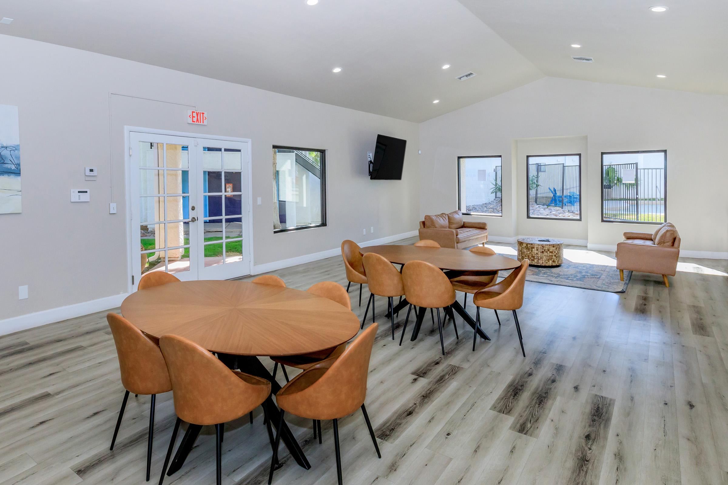 Interior of a modern community room with a large round wooden table surrounded by brown chairs. There's a cozy seating area with a couch and a rug, large windows providing natural light, and a TV mounted on the wall. The space has neutral-colored walls and a hardwood floor, creating a welcoming atmosphere.