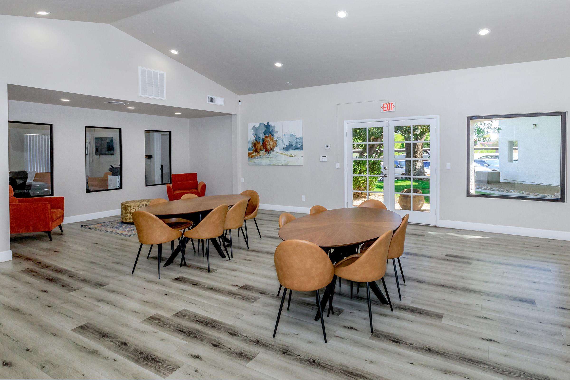 A modern interior space featuring wooden floors, multiple round tables with tan chairs, and a bright, airy atmosphere. There are large windows allowing natural light, along with a cozy seating area and decorative wall art, creating a welcoming environment.