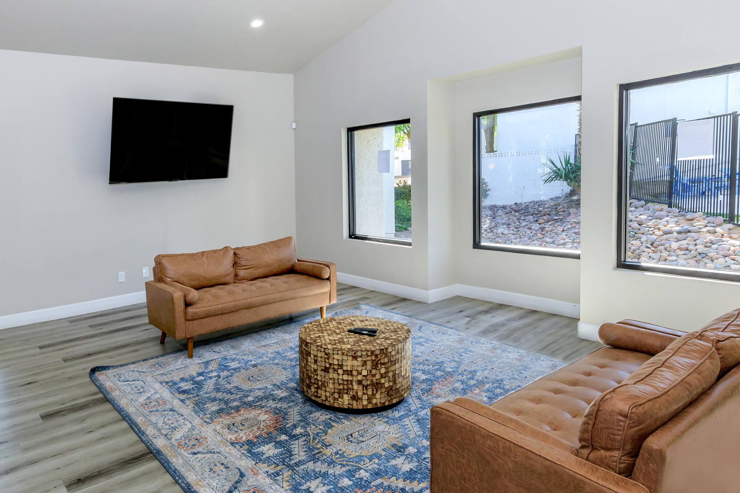 A contemporary living room featuring two brown leather sofas, a patterned rug on wooden flooring, and a circular coffee table. Large windows allow natural light to flood the space, highlighting the minimalist design and modern decor. A flat-screen TV is mounted on the wall.