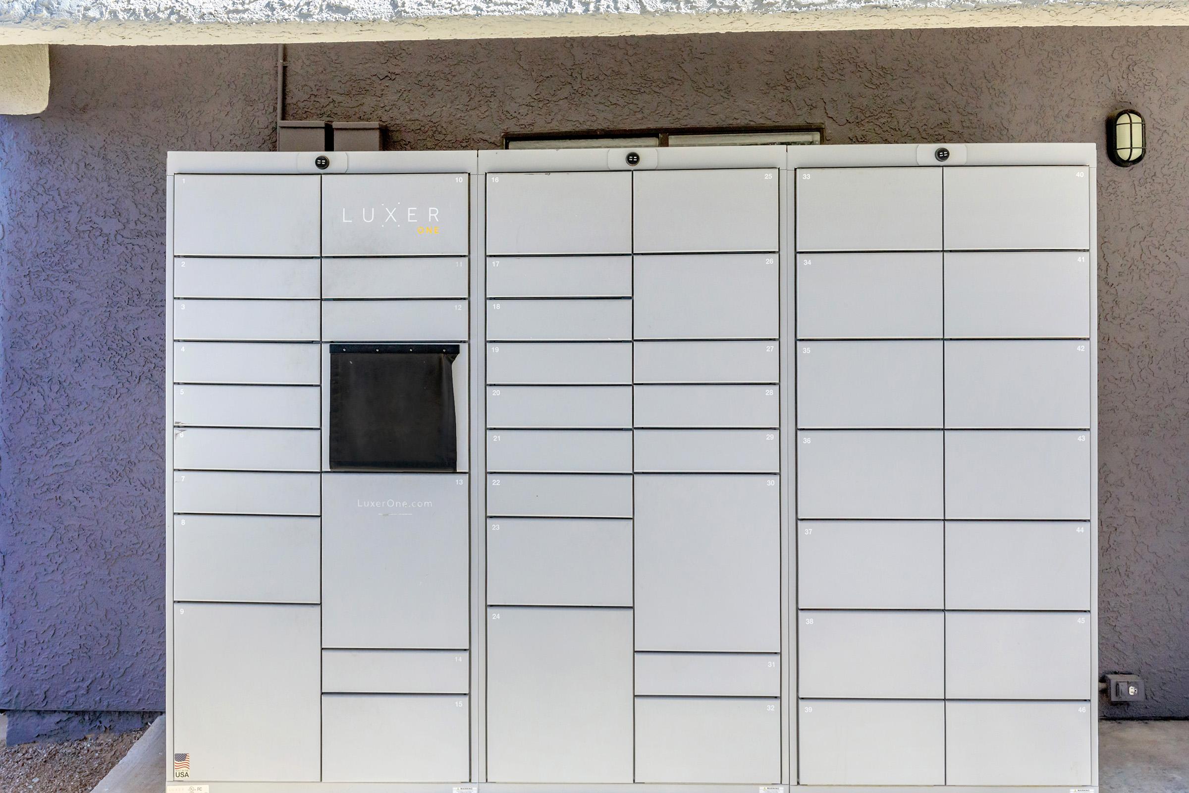 A gray package delivery locker system with multiple compartments, labeled “LUXER.” The lockers are installed against a textured wall, with a light fixture mounted above. The locker section on the far left has a black access panel.