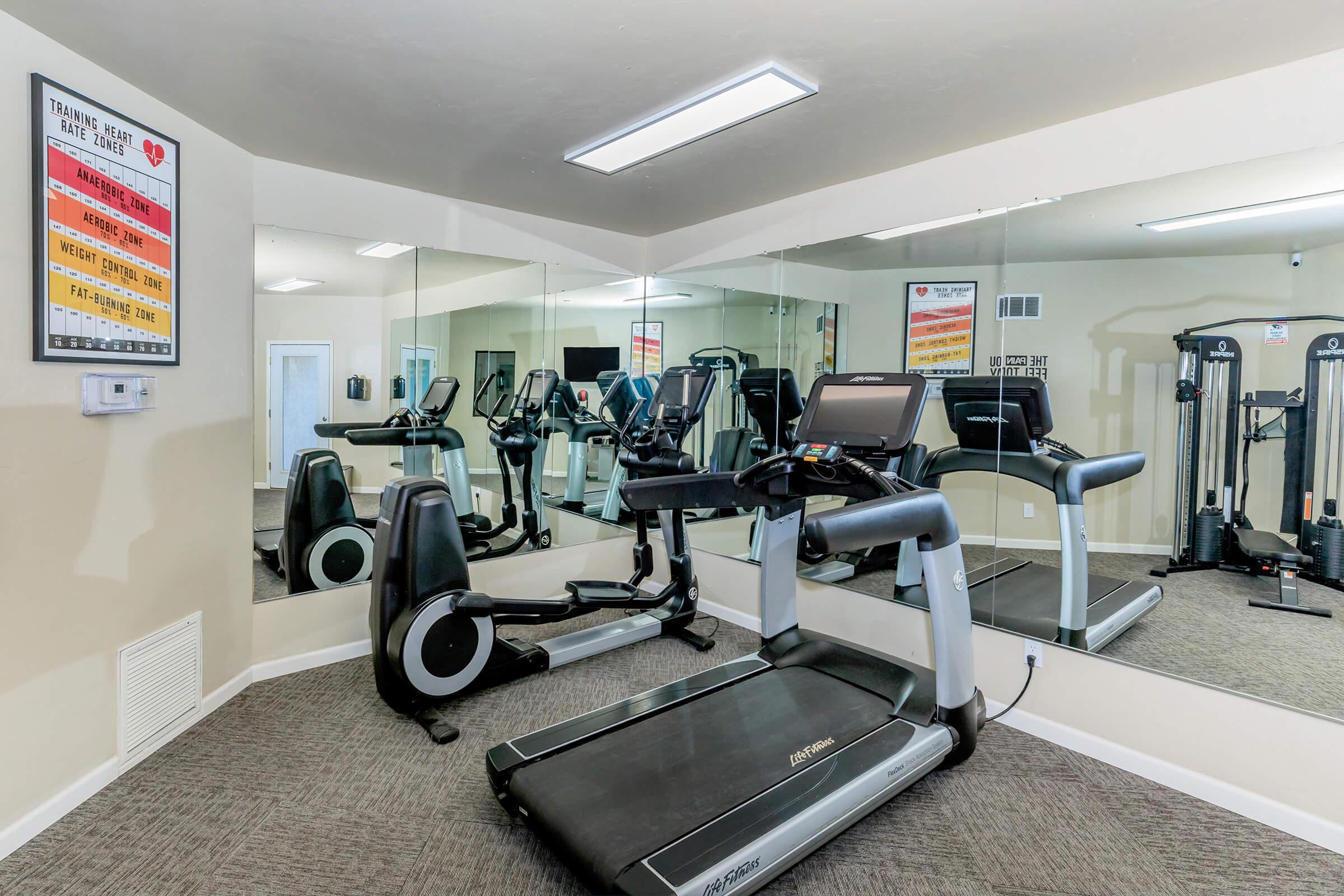 A fitness room featuring a treadmill in the foreground and various exercise equipment in the background. Wall mirrors reflect the gym space, and motivational posters are displayed on the walls. The flooring is carpeted, creating a welcoming workout environment.