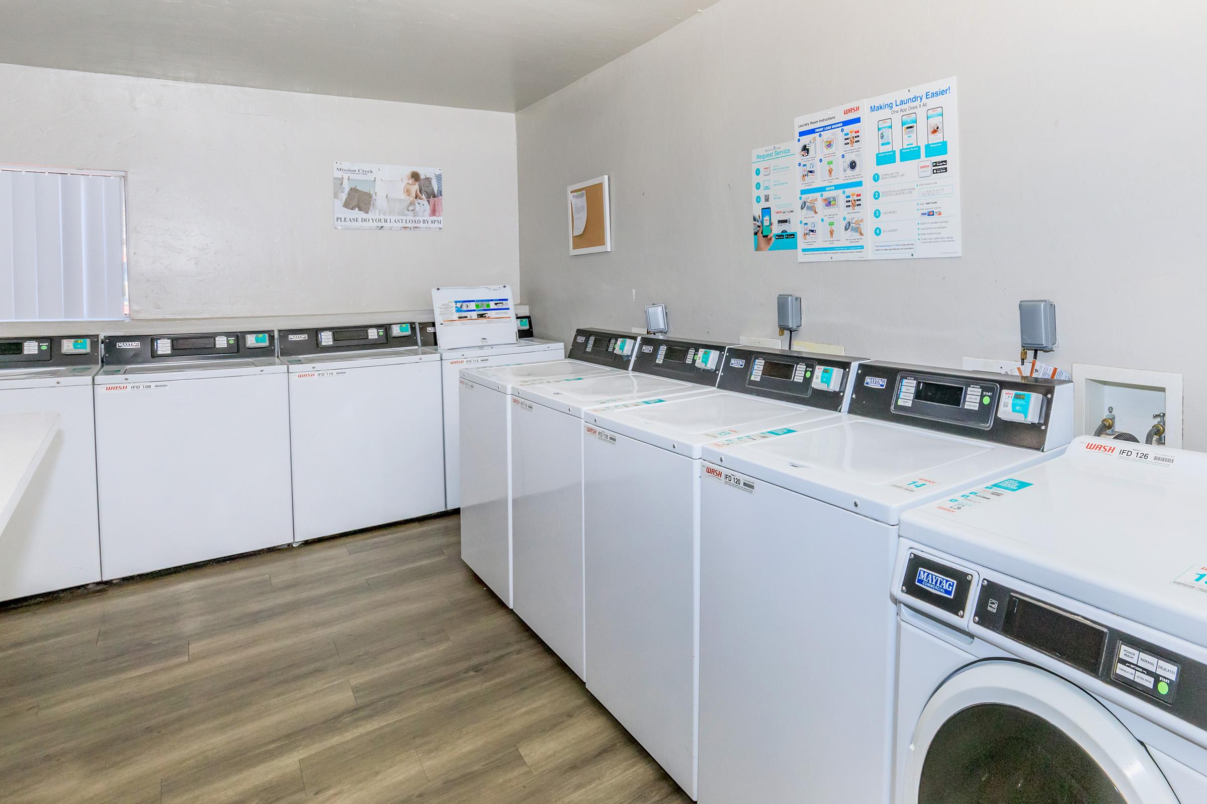 A modern laundry room featuring several white washing machines and dryers lined up against the wall. The space is brightly lit, with informational posters on the walls and a clean, minimalist design. The floor is made of light-colored wood, creating a neat and organized atmosphere.