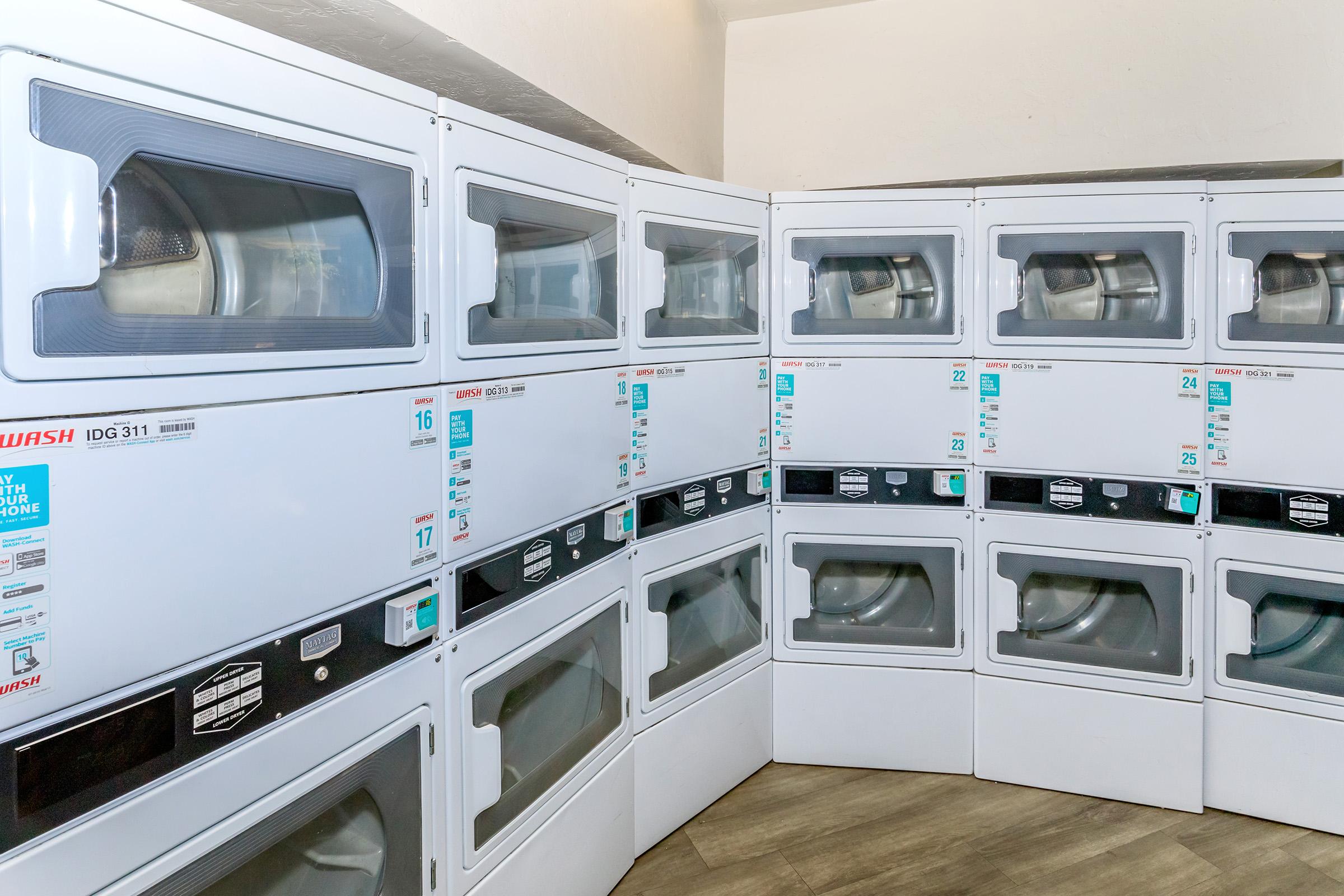 A row of stacked commercial dryers in a laundry facility, featuring digital displays and various settings. The dryers are arranged against a wall, providing a clean and organized appearance, suitable for public use.