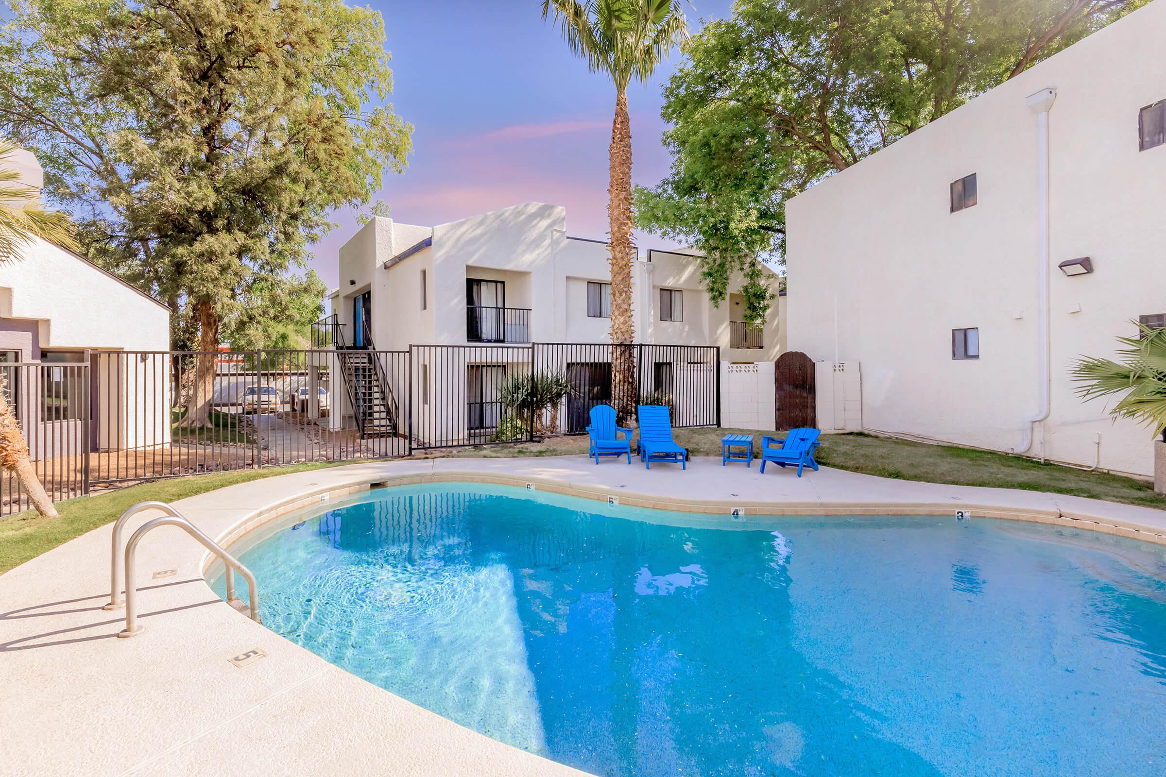 A tranquil outdoor swimming pool surrounded by palm trees and blue lounge chairs. In the background, there are white apartment buildings and a fenced area. The sky has a soft pastel hue, suggesting a serene evening or morning atmosphere.