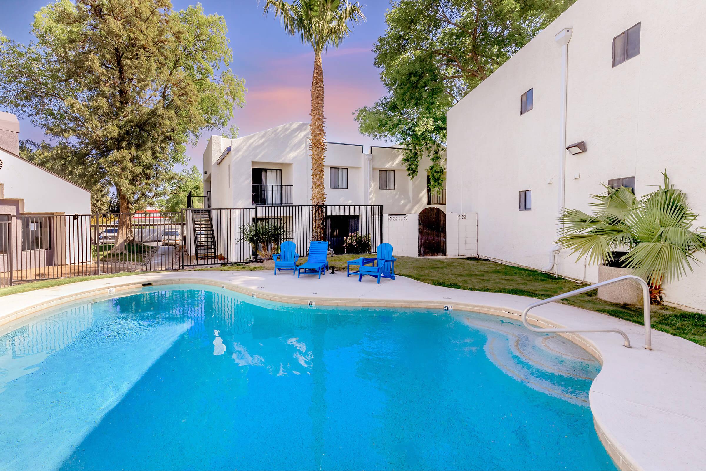 A serene outdoor swimming pool surrounded by lounge chairs and lush greenery. The area features a tall palm tree, white buildings in the background, and a well-maintained lawn, creating a relaxing atmosphere ideal for leisure and sunbathing.