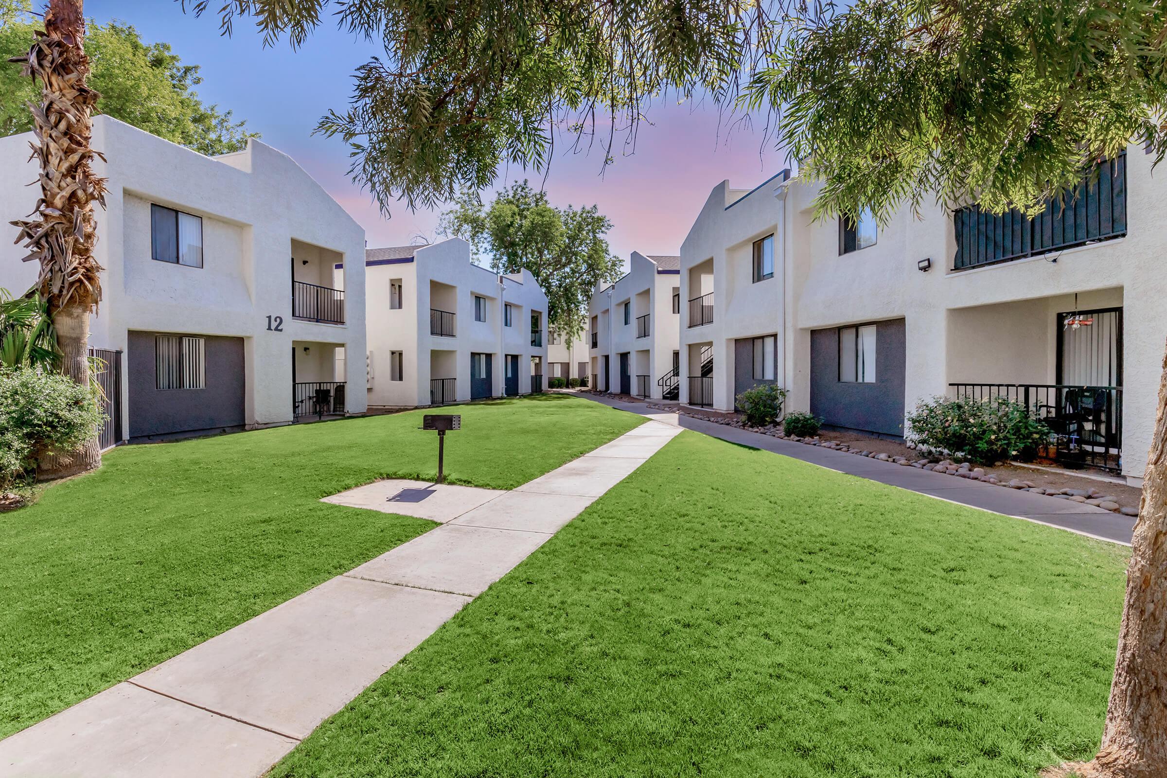 A view of a modern apartment complex showcasing two-story buildings arranged around a well-maintained green lawn. A pathway connects the units, with palm trees and shrubs enhancing the landscaping. The sky above features soft pastel colors, indicating a pleasant day.
