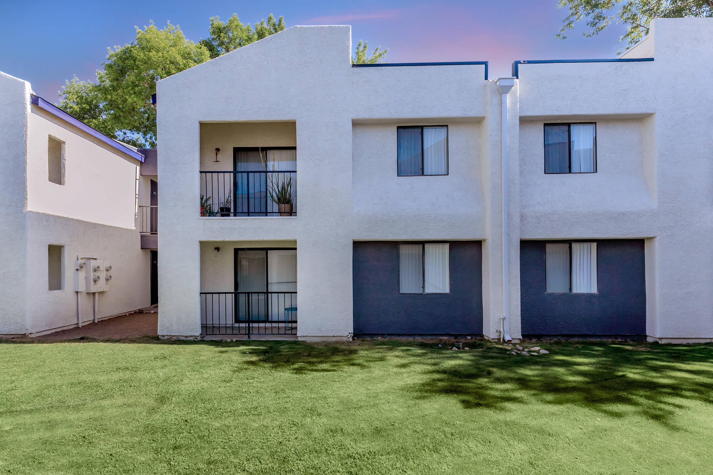 Two-story apartment building featuring a white exterior with gray accents. Each unit has a small balcony with black railings, and the ground is covered with green grass. Trees are visible in the background, providing a natural setting.