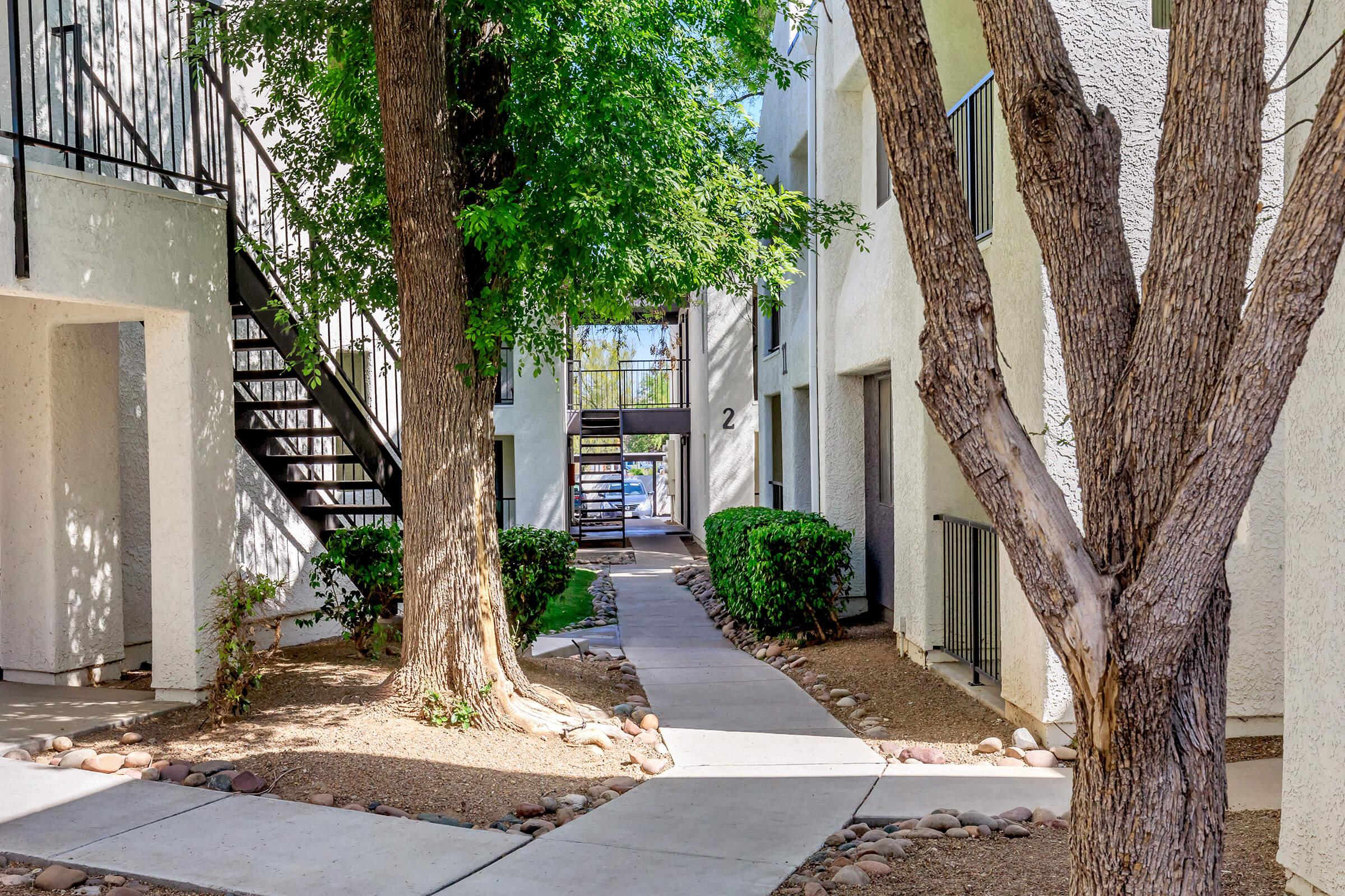 A peaceful apartment complex courtyard featuring a paved walkway lined with small rocks, surrounded by manicured bushes and trees. A set of stairs leads to upper-level apartments, and a glimpse of a pool area is visible in the background through the archway.