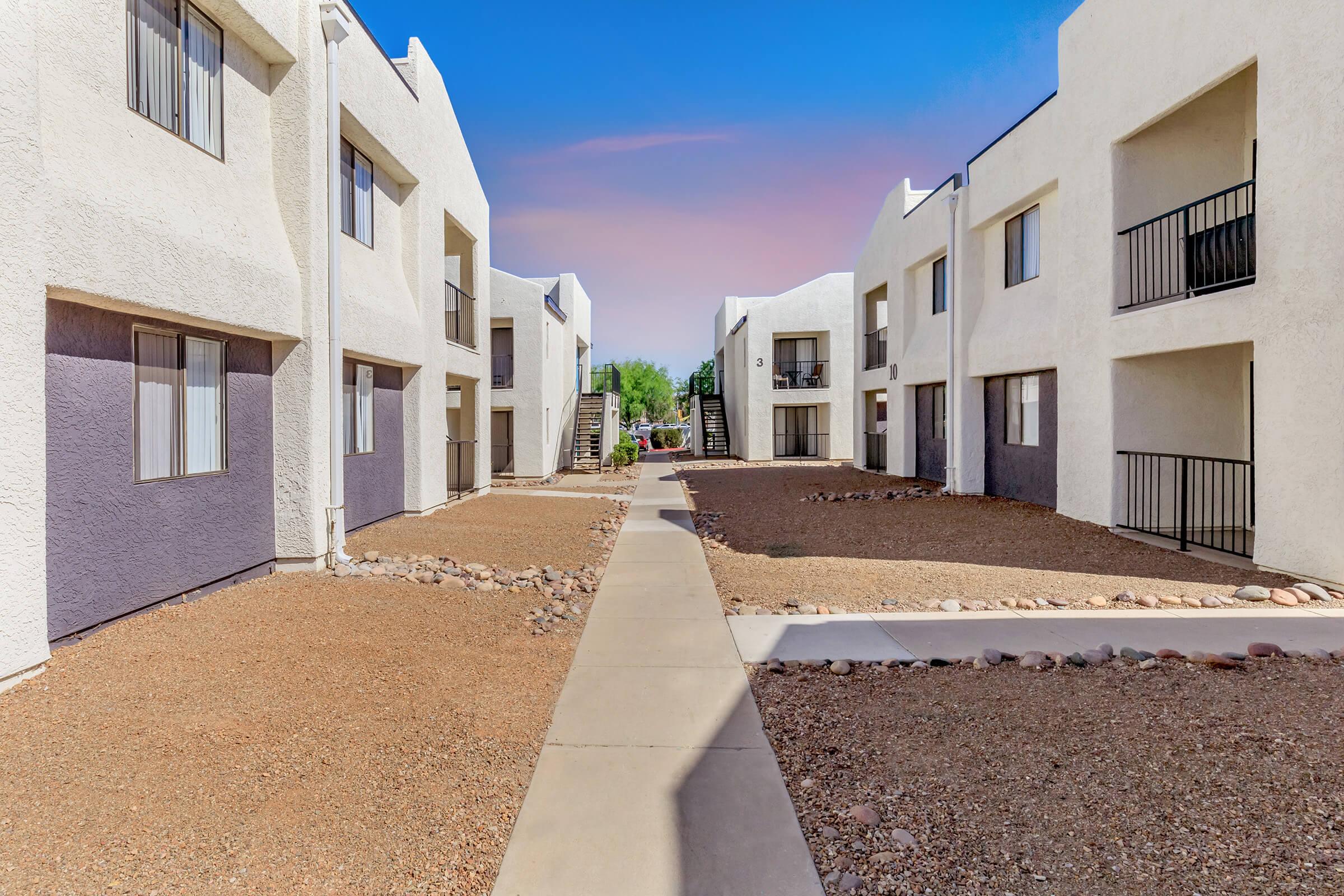 View of a modern apartment complex with two-story buildings featuring white exteriors. A central pathway runs through the courtyard, surrounded by gravel landscaping and small rocks. Balconies are visible on the upper floors, and clear blue skies provide a bright ambiance.