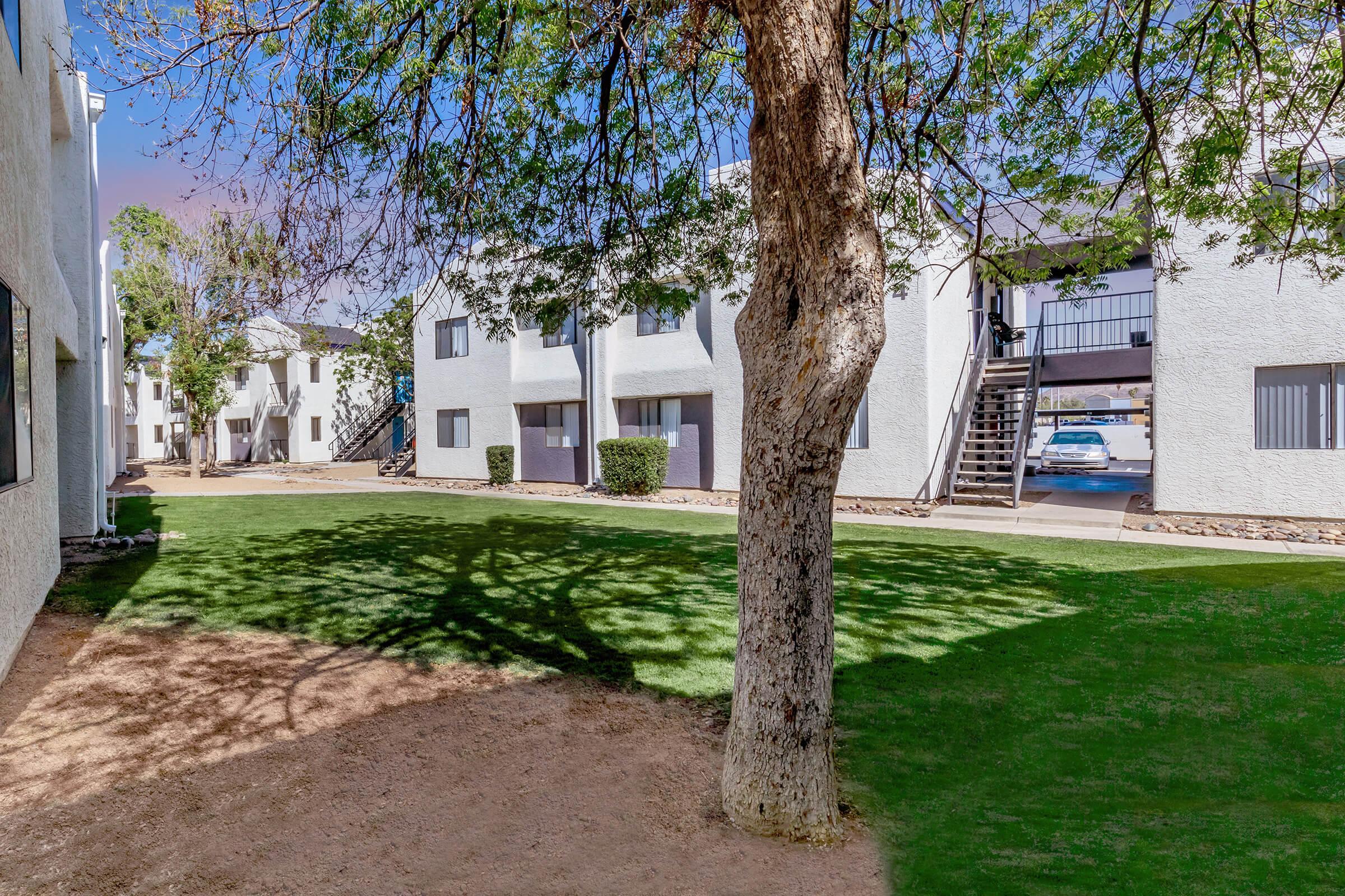 A courtyard with green grass surrounded by white apartment buildings. A tree is in the foreground casting shadows on the ground, and there are stairs leading to upper levels of the building. In the background, a parking area is visible with a parked blue car. The sky is clear and bright.