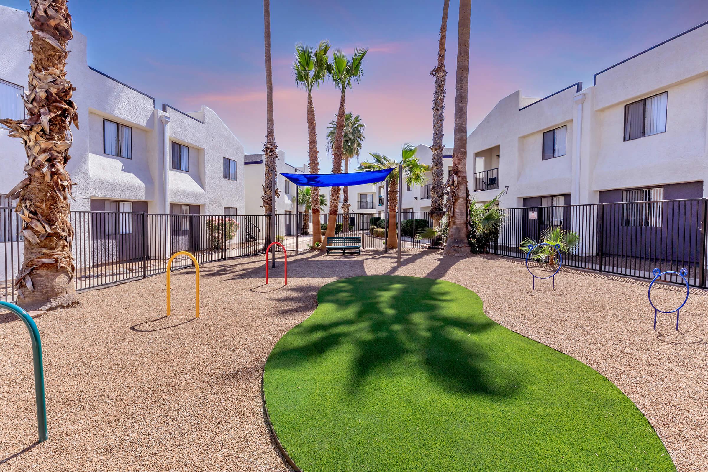 A landscaped courtyard in an apartment complex featuring a green lawn area, palm trees, and a blue shade structure. Play equipment with colorful arches is visible, surrounded by gravel and fencing. The background shows white apartment buildings under a colorful sky.