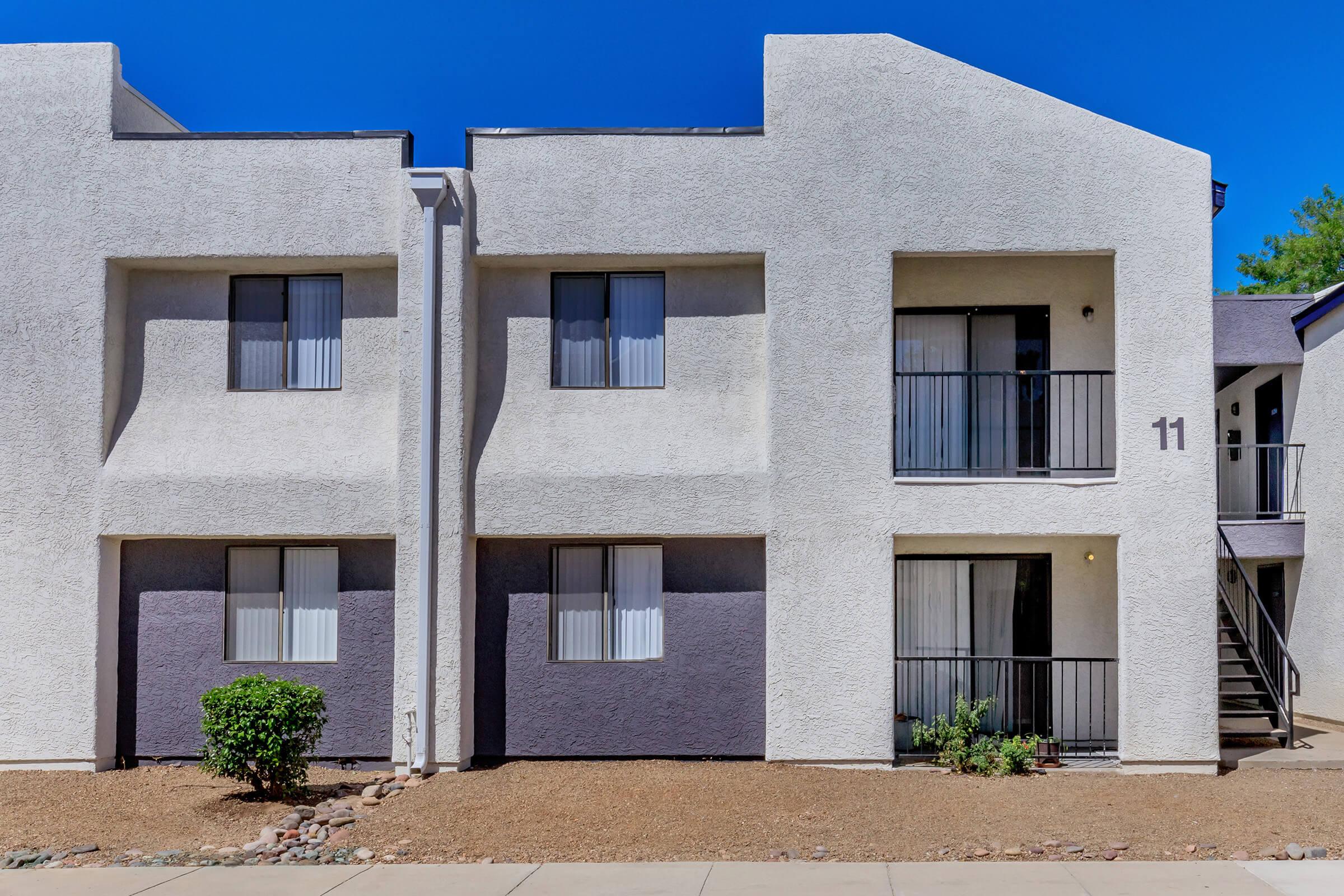 Two-story apartment building with a modern design, featuring light-colored stucco walls and large windows covered by sheer curtains. The building has contrasting dark gray accents, a small plant in front, and a paved walkway. The unit number "11" is visible on the second floor. Clear blue sky in the background.