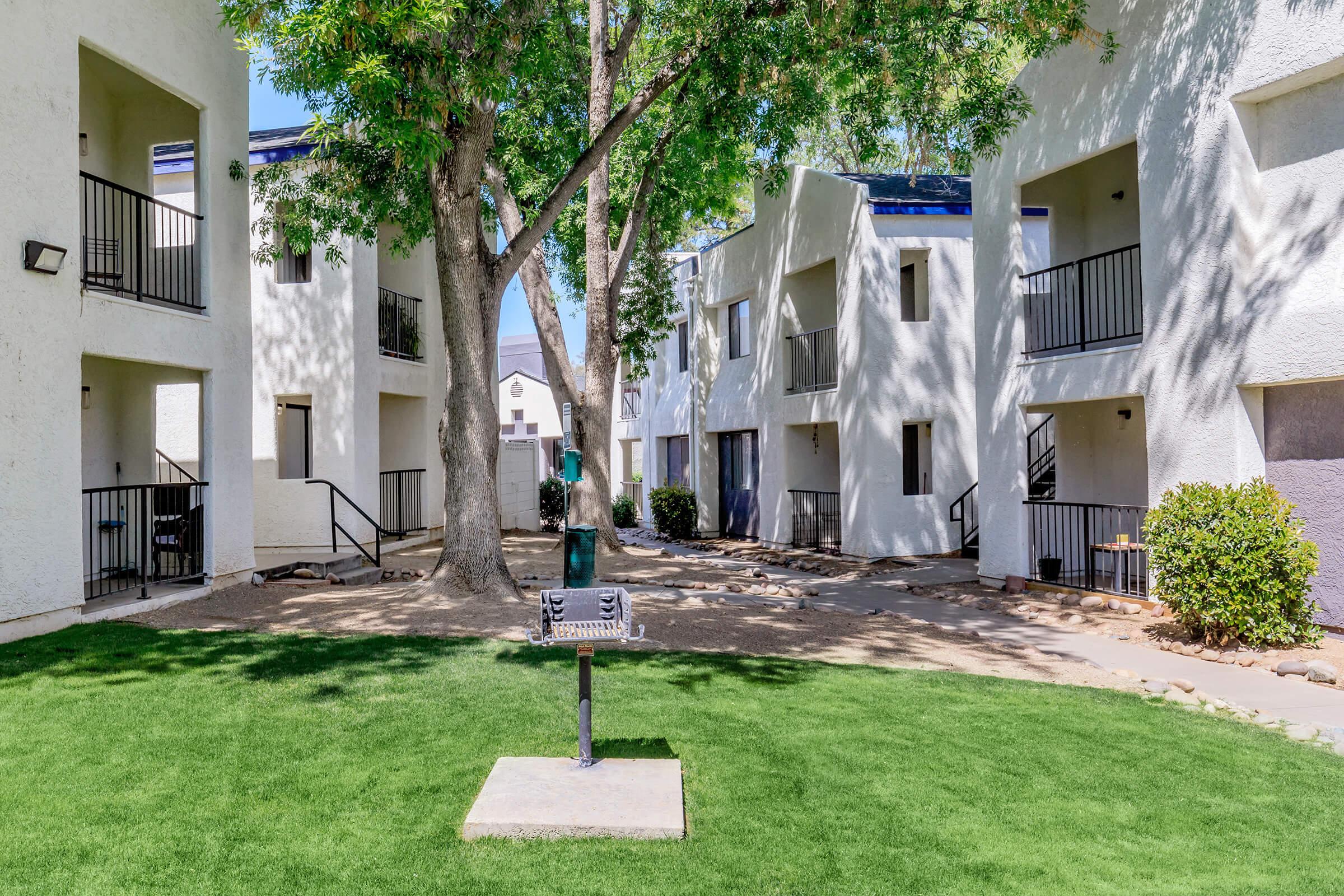 View of a landscaped courtyard featuring green grass, tall trees, and two white apartment buildings with balconies. A barbecue grill is positioned on the lawn, creating an inviting outdoor space. The area is bright with natural sunlight filtering through the trees.