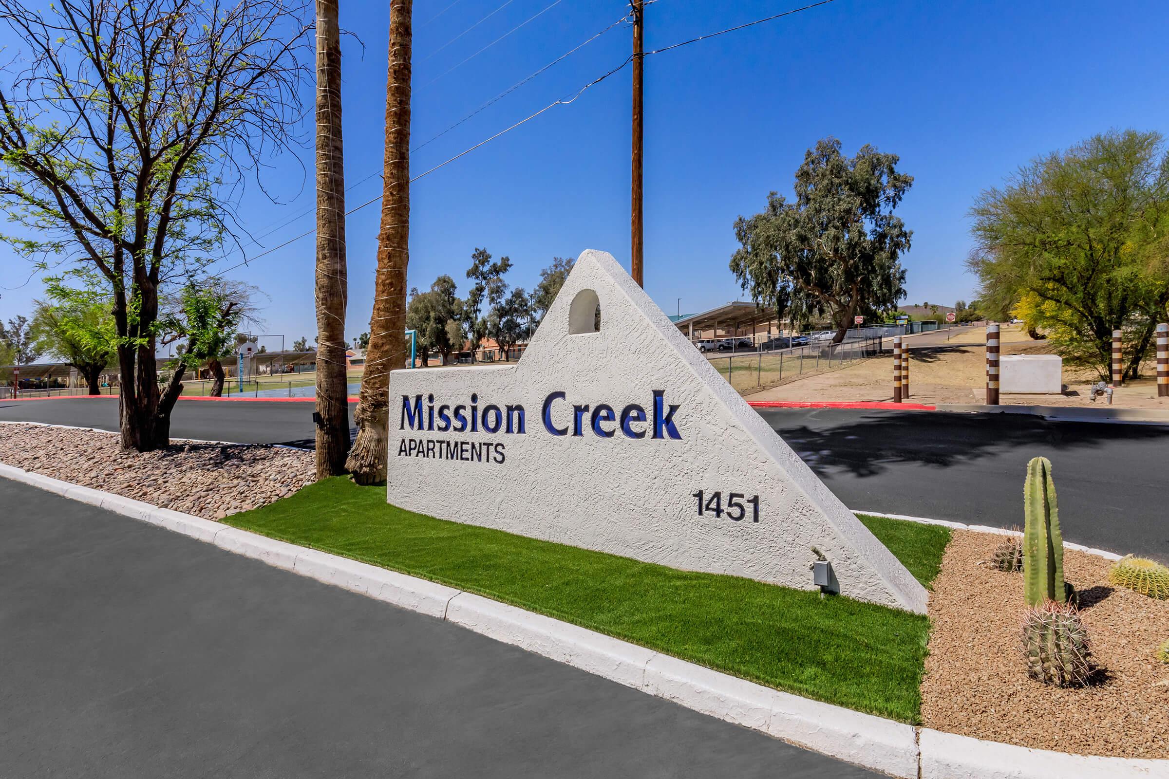 Sign for Mission Creek Apartments located at 1451, featuring a triangular white structure with blue lettering. Surrounding landscape includes desert vegetation, palm trees, and a well-maintained road, set against a clear blue sky. The area appears sunny and inviting.