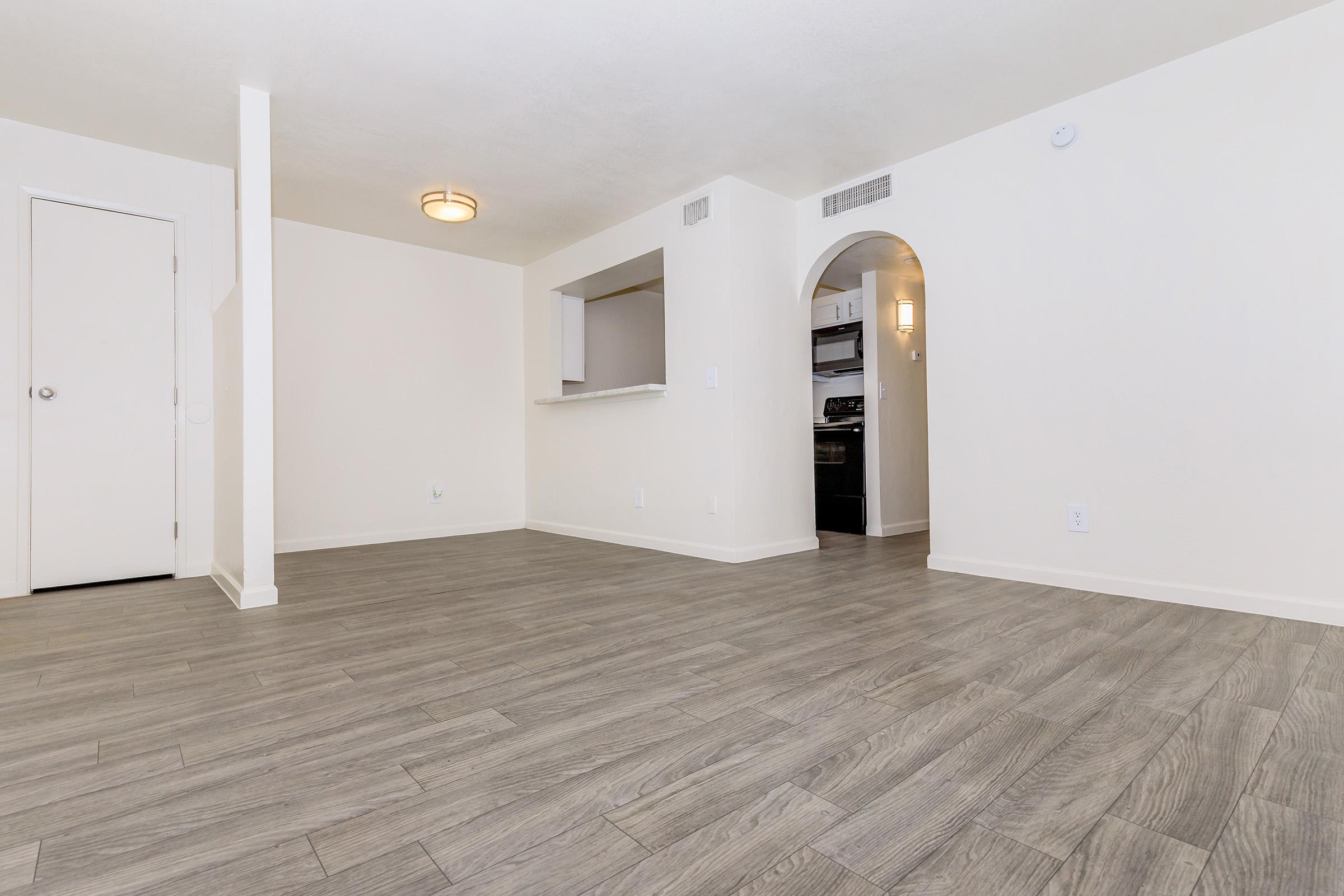 An empty, well-lit room with light-colored walls and wooden-like flooring. An archway leads to a kitchen area visible in the background. A door is on the left, and a ceiling fixture is above. The space appears modern and inviting.