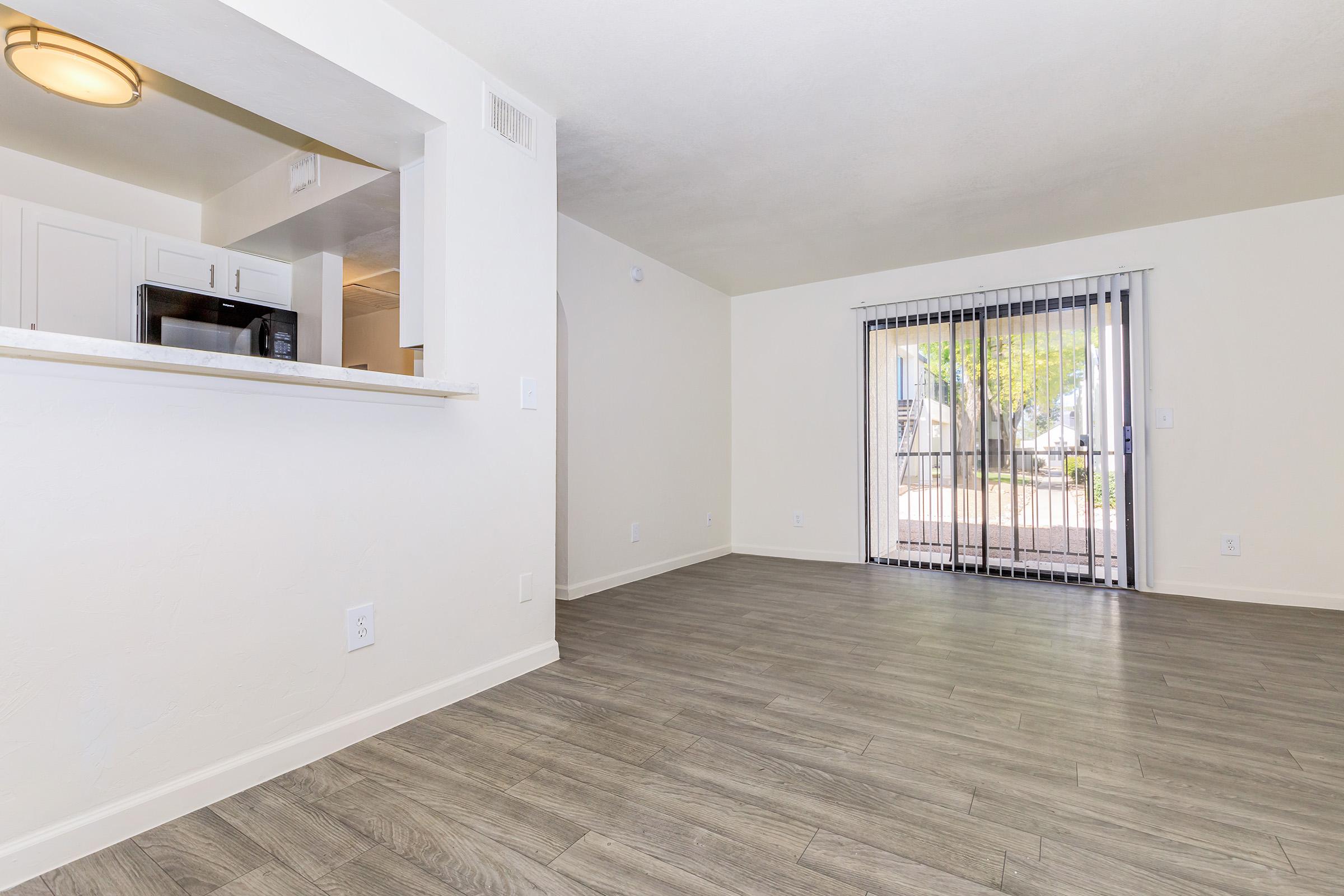 Interior view of a bright, modern living space featuring light-colored walls, laminate flooring, and a sliding glass door leading to an outdoor area. A kitchen area is visible in the background, with white cabinetry and a microwave. Natural light illuminates the room, creating an inviting atmosphere.
