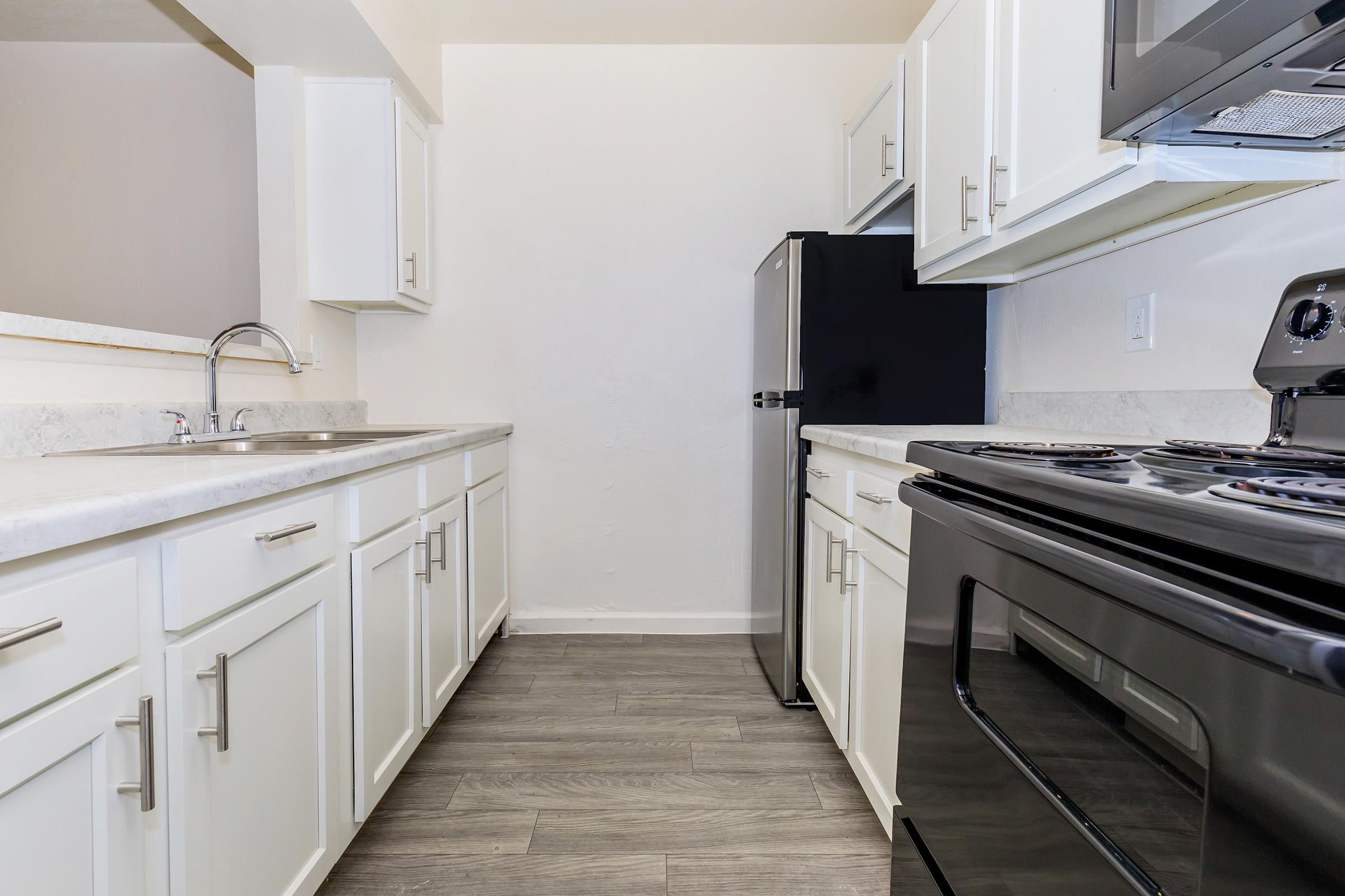 A modern kitchen featuring white cabinets, a granite countertop, stainless steel sink, and a black refrigerator and stove. The space is well-lit, showcasing a clean and organized design with laminate flooring, creating an inviting atmosphere.