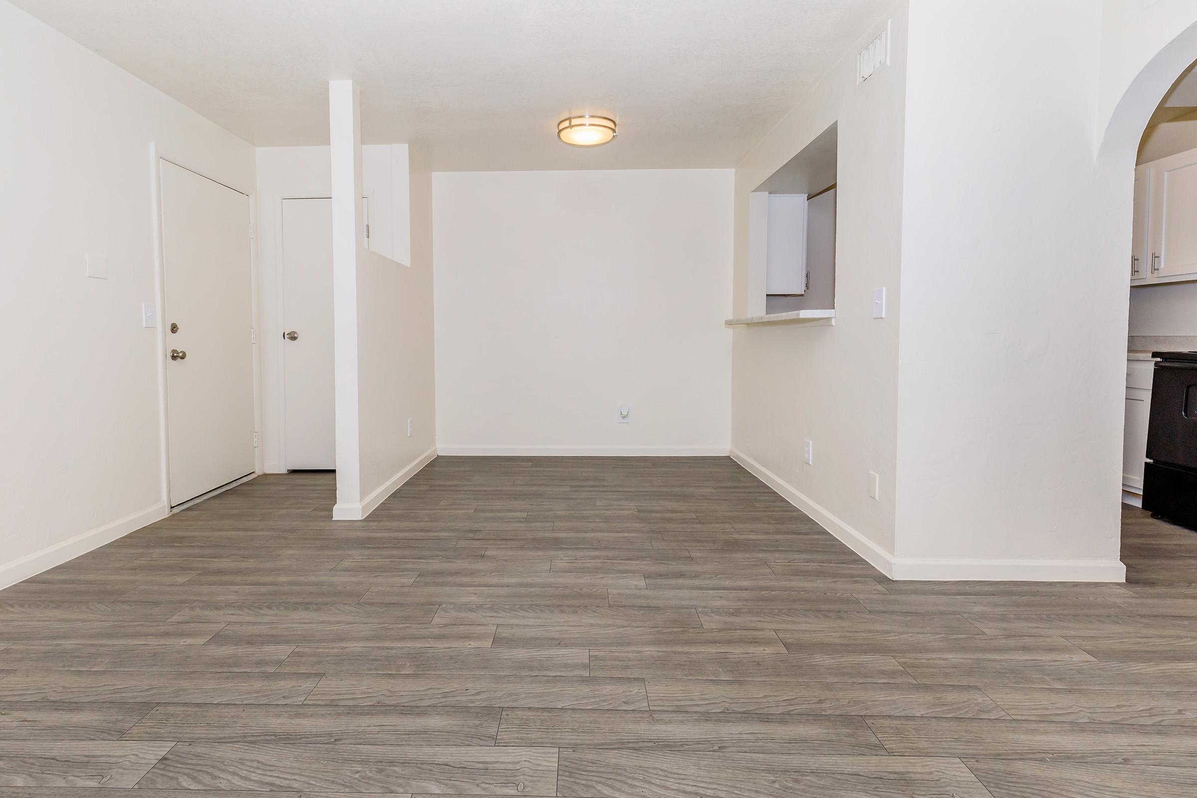 Spacious, empty living area with light-colored walls and gray laminate flooring. A kitchen area is visible in the background with a white countertop and black appliances. A doorway leads to an entrance, and the ceiling features a round light fixture. The overall atmosphere is bright and minimalistic.