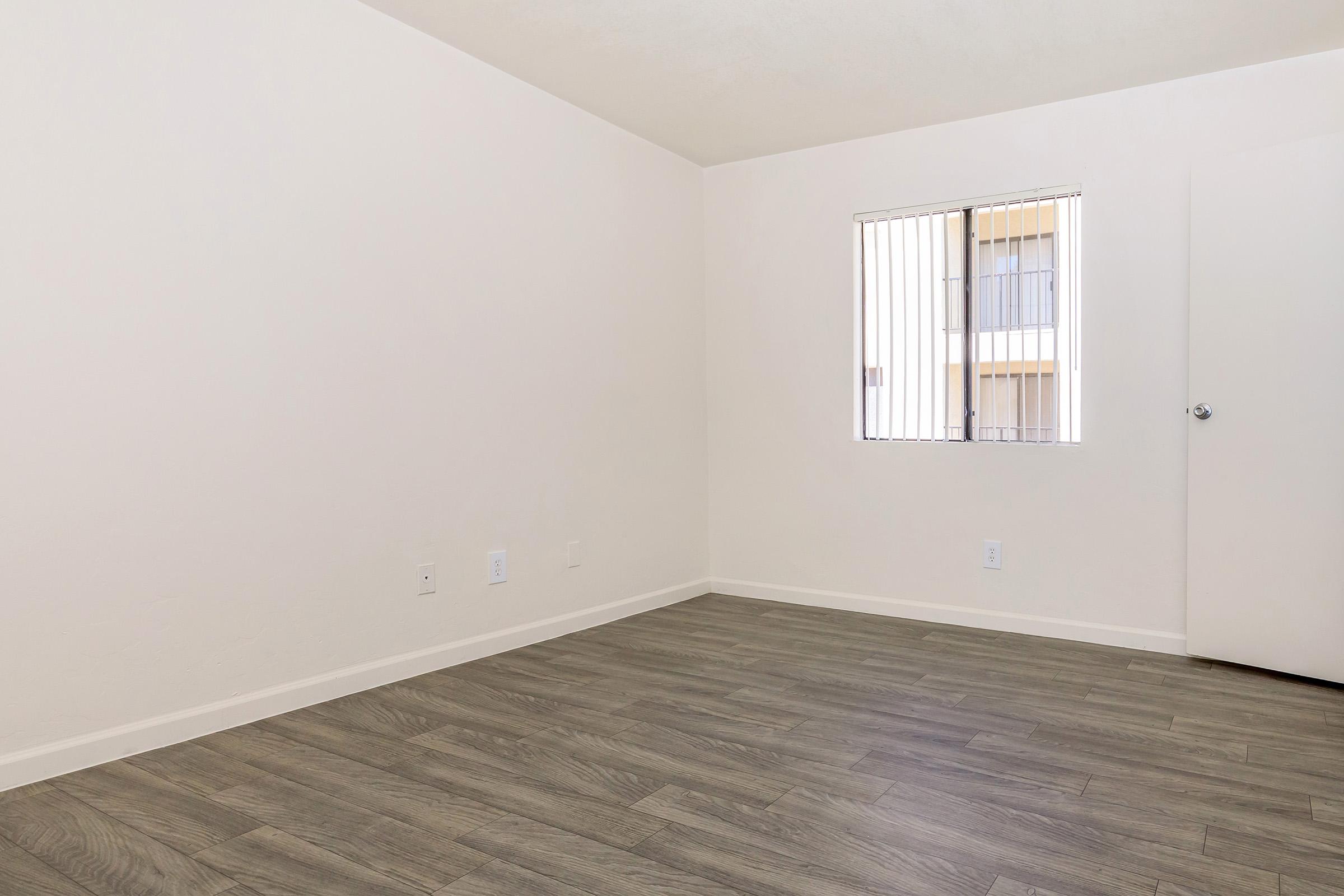 A vacant room featuring light-colored walls and a wooden floor. There is a single window with vertical blinds allowing natural light, and a door on the right side. The space is empty, creating a minimalist atmosphere.