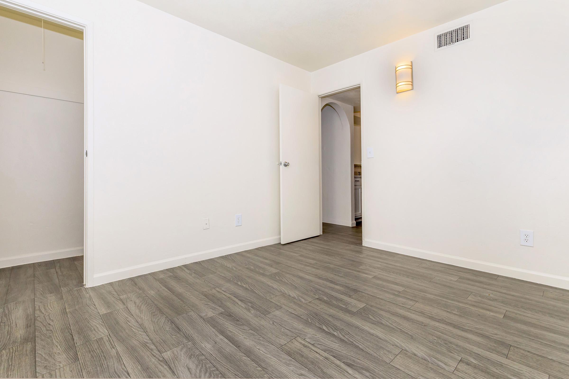 An empty room featuring light-colored walls and a smooth wooden floor. There is a door leading to another room and an open closet space on the left. A wall sconce provides soft lighting, and the overall ambiance is bright and spacious.