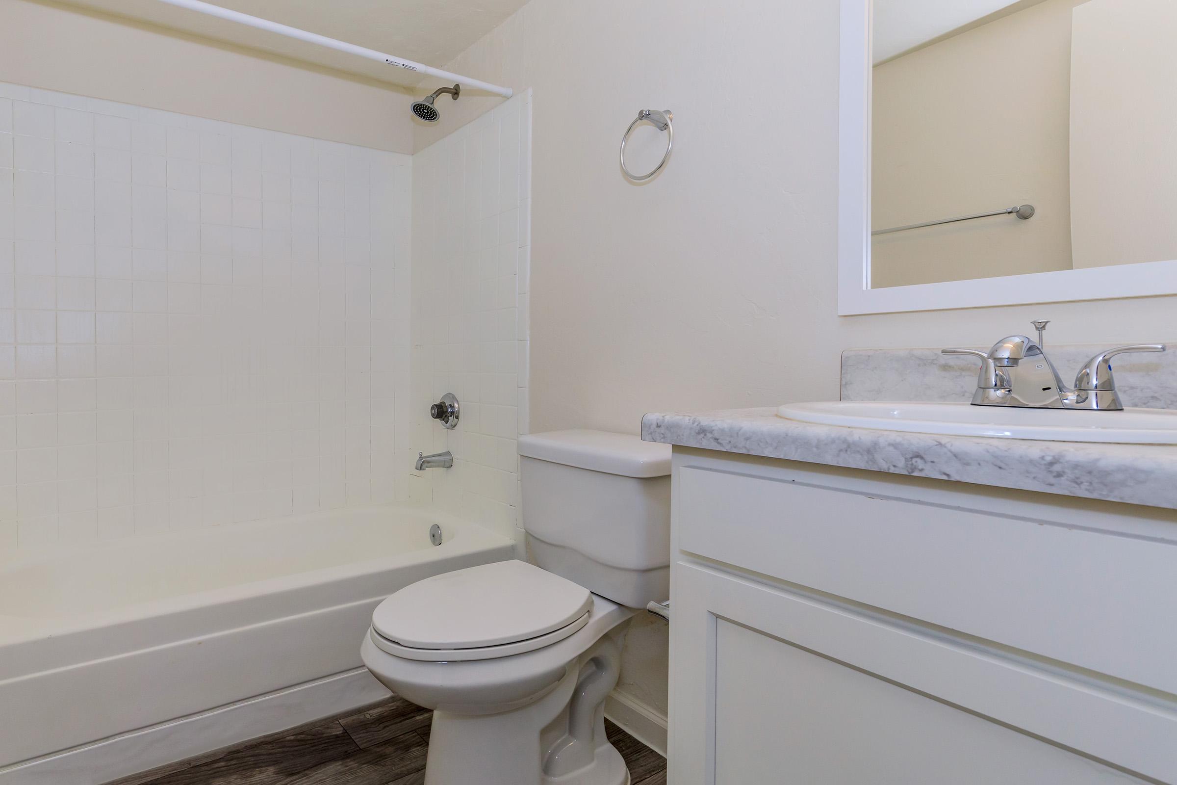 A clean, modern bathroom featuring a white bathtub with a shower, a sleek toilet, and a vanity with a marble countertop and mirror. The walls are painted a light color, and there’s a towel holder on the wall, creating a bright and simple aesthetic.