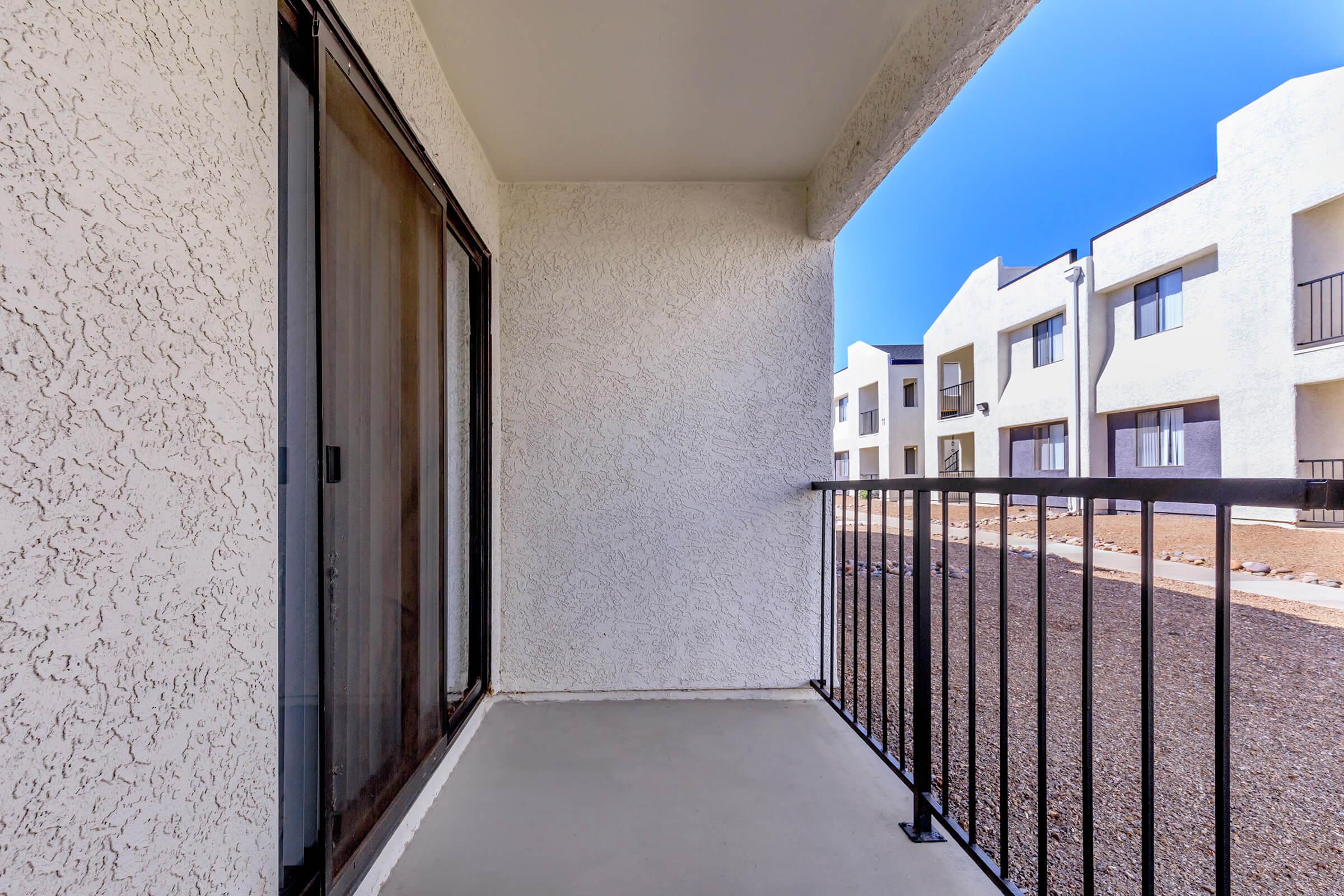 A view from a balcony featuring a textured white wall and a black railing, overlooking a courtyard with several white buildings. The sky is clear and blue, and the ground is covered with gravel, creating a simple outdoor space.