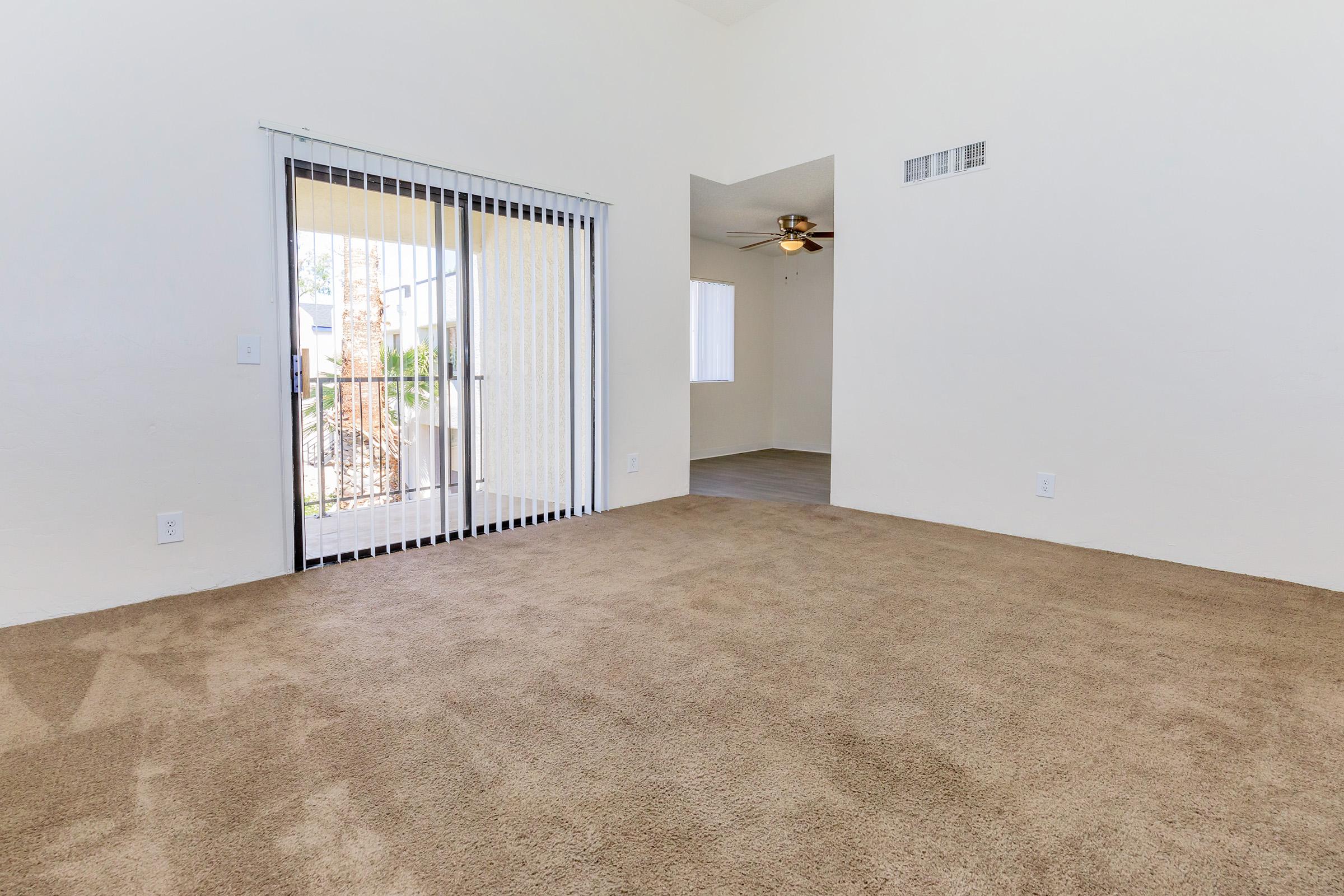 A spacious room featuring beige carpet flooring, sliding glass doors leading to an outdoor area, and a doorway to another room with a ceiling fan. The walls are painted white, creating a bright and open atmosphere.