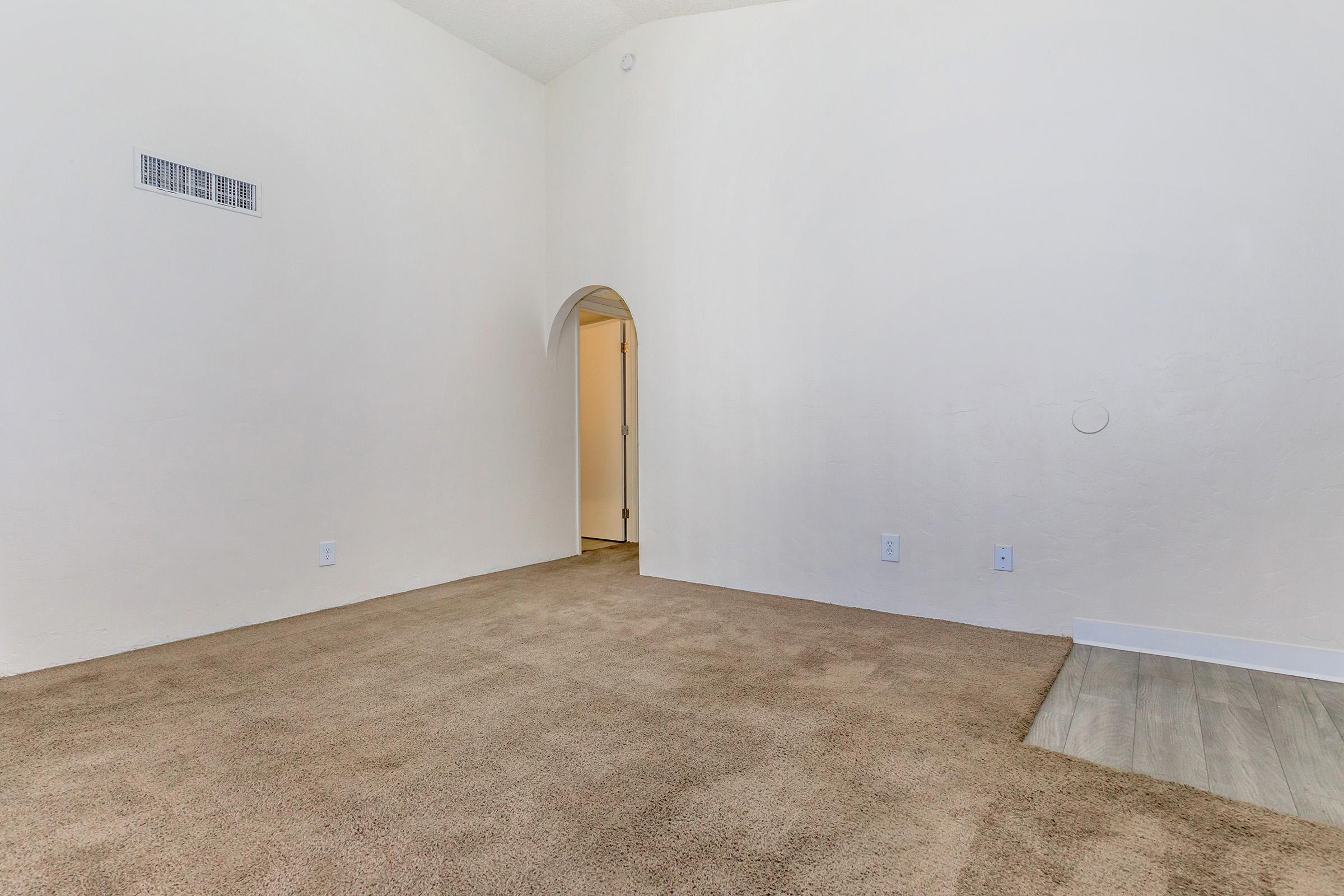 A spacious, empty room with light beige carpet, white walls, and a small archway leading to another room. There is a vent near the ceiling and a couple of small light switches on the walls. The floor transitions to a different surface near the archway. Natural light enters the space, creating a bright atmosphere.
