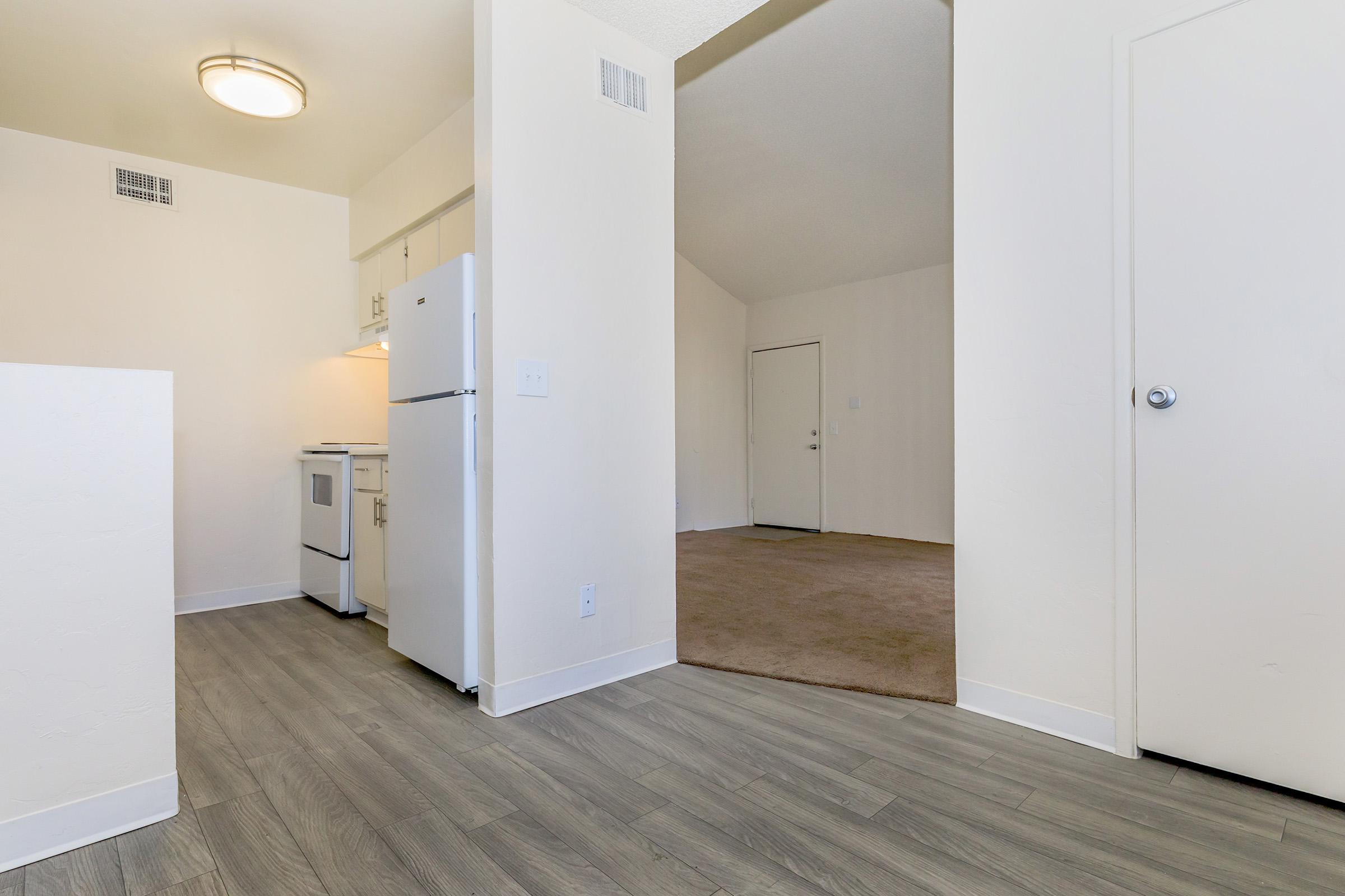Interior view of an open apartment space featuring a kitchen area with a refrigerator and stove on the left, light-colored walls, and wooden flooring. A doorway leads to a carpeted living area, creating a spacious layout. Well-lit with a ceiling light fixture visible in the kitchen.