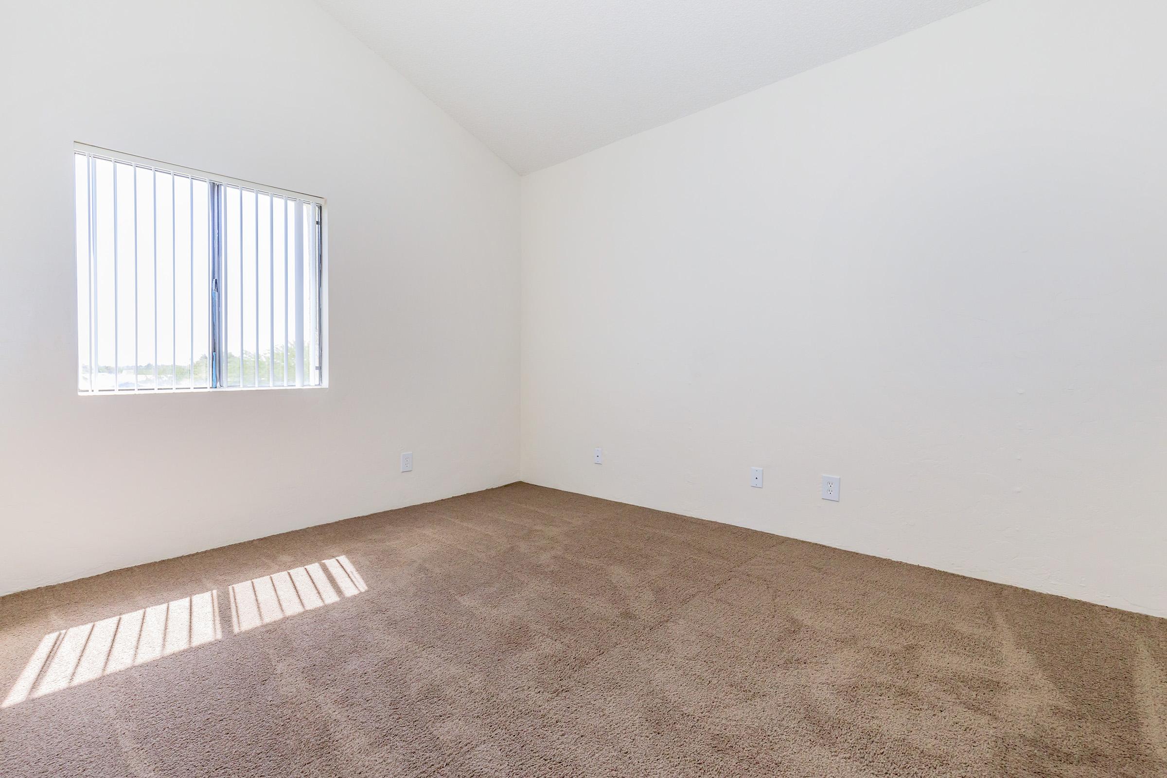 A vacant room with light beige walls and a high ceiling. The floor is covered in light brown carpet. A window with vertical blinds allows natural light to enter, creating a bright atmosphere. The room appears spacious and unfurnished.