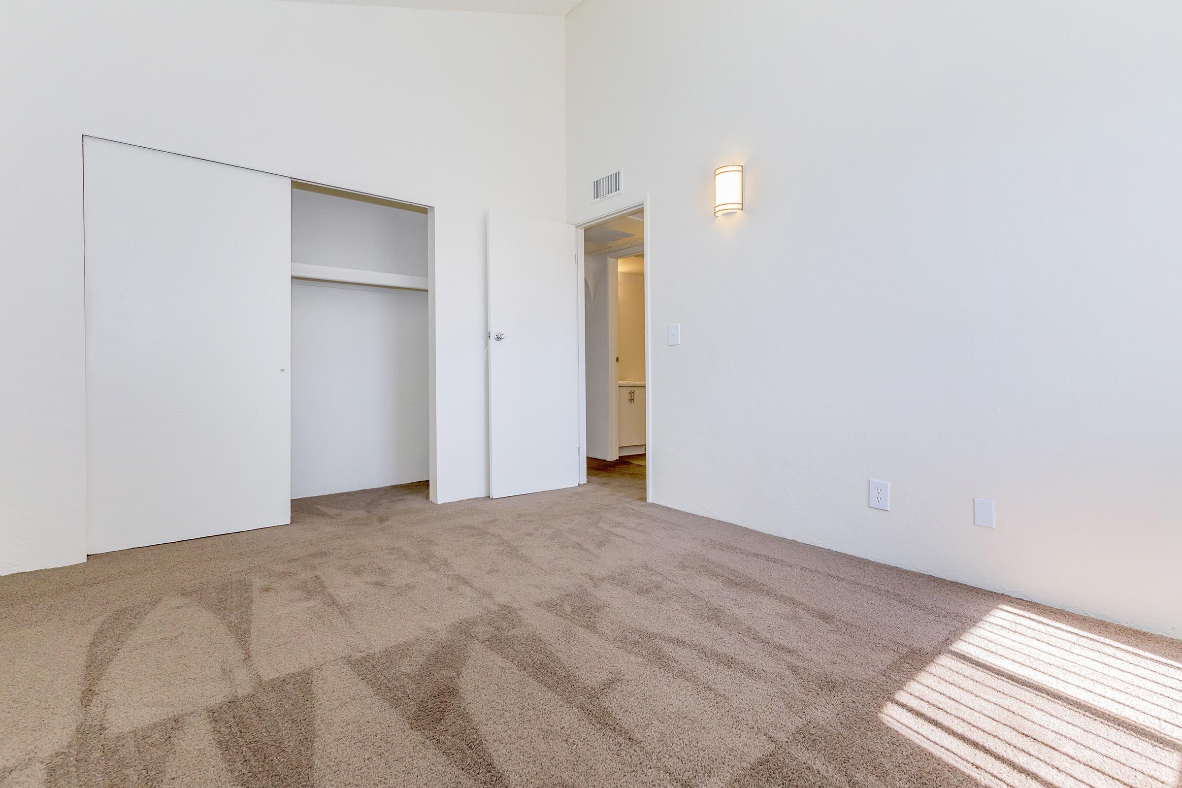 A spacious, empty room with light beige carpet and white walls. There are two doorways: one leading to a closet and another to a bathroom. A wall-mounted light fixture illuminates the space, and natural light casts soft shadows on the floor. The overall atmosphere is bright and inviting.