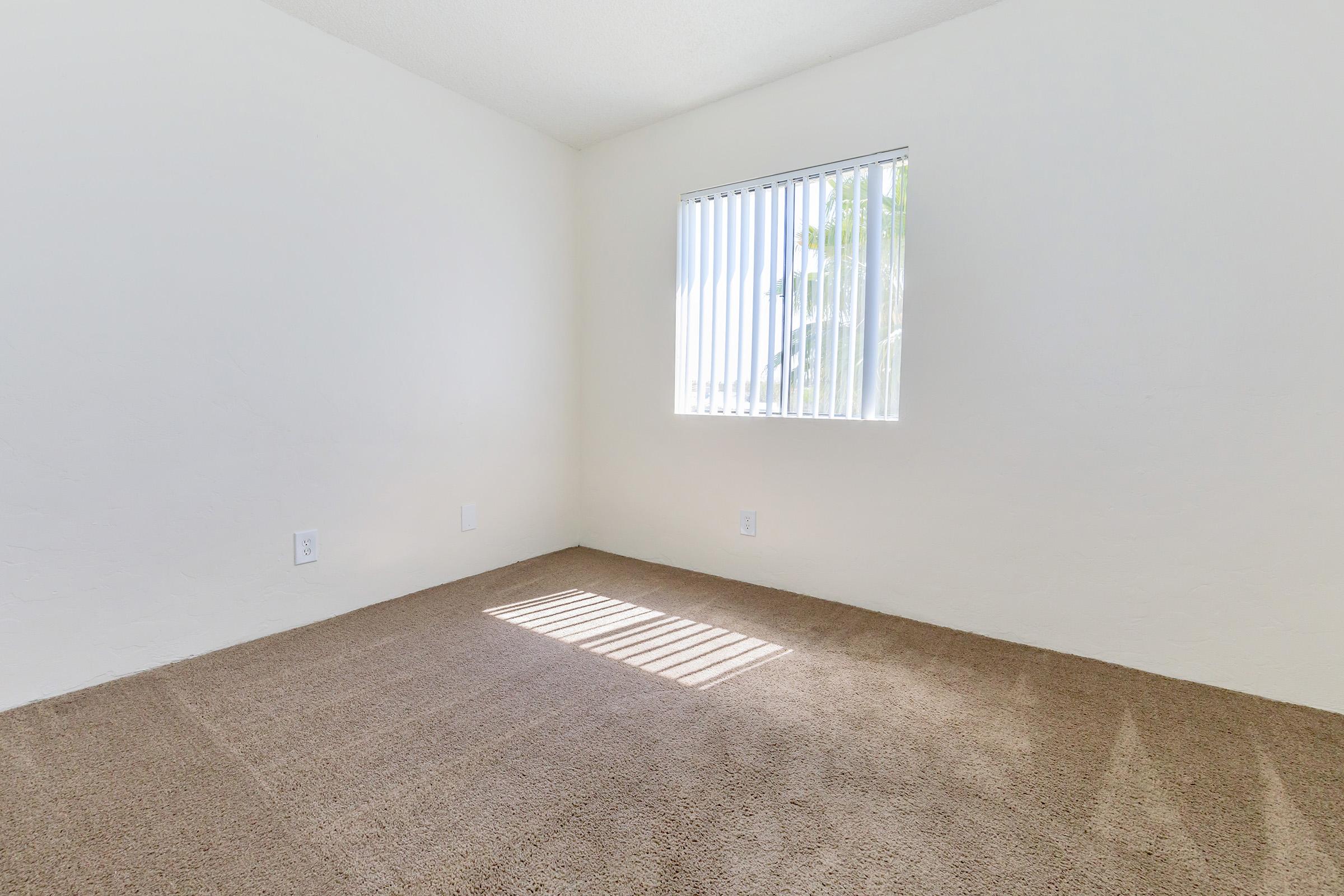 Empty room with light beige carpet and white walls. A window with vertical blinds allows sunlight to stream in, creating a striped pattern on the floor. The space is uncluttered, with no furniture, giving it a bright and open feel.