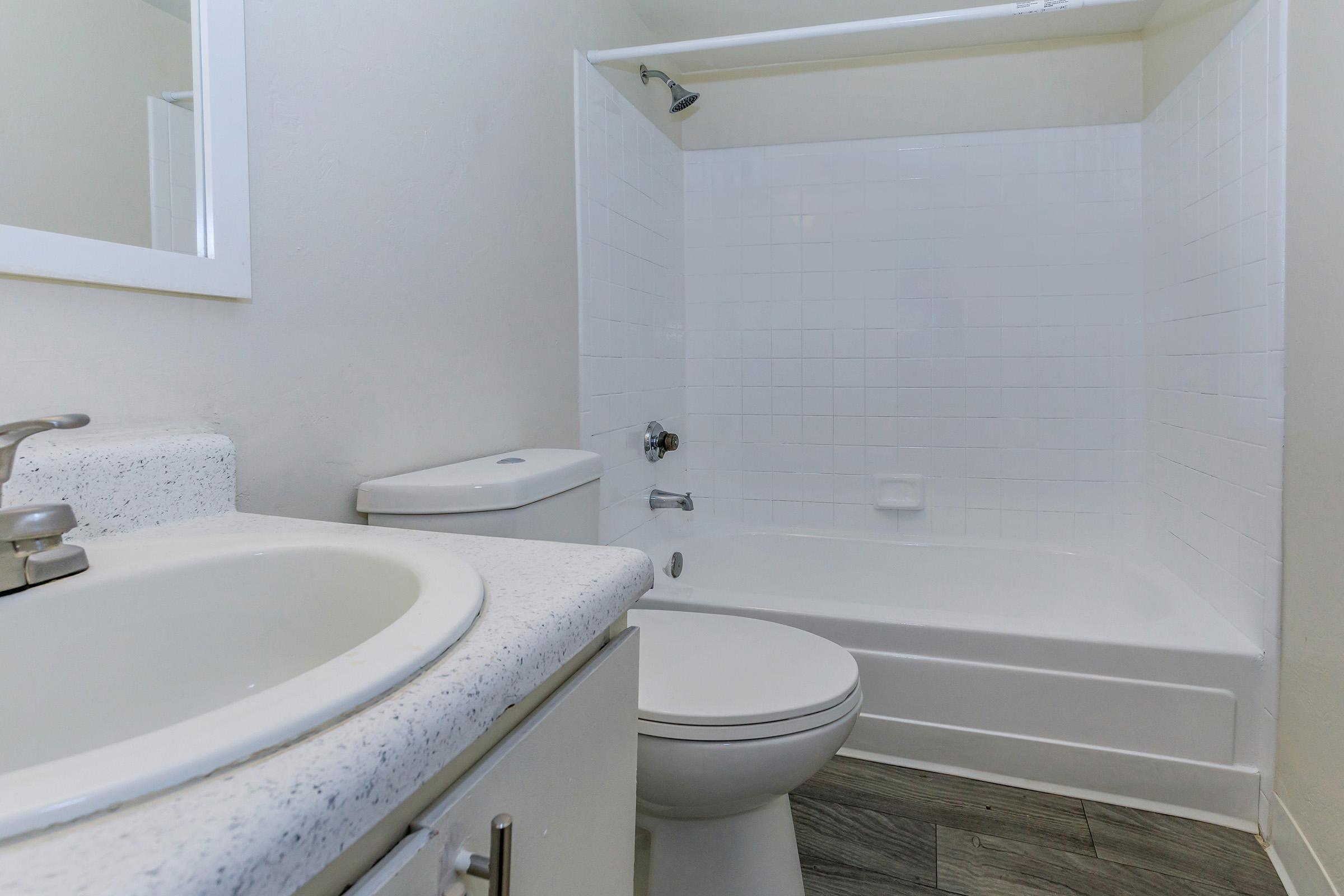 A clean, modern bathroom featuring a white bathtub with a showerhead, a white toilet, and a sink with a speckled countertop. The walls are painted a light color, and the floor is tiled. A mirror is mounted above the sink, completing the simple, functional design.
