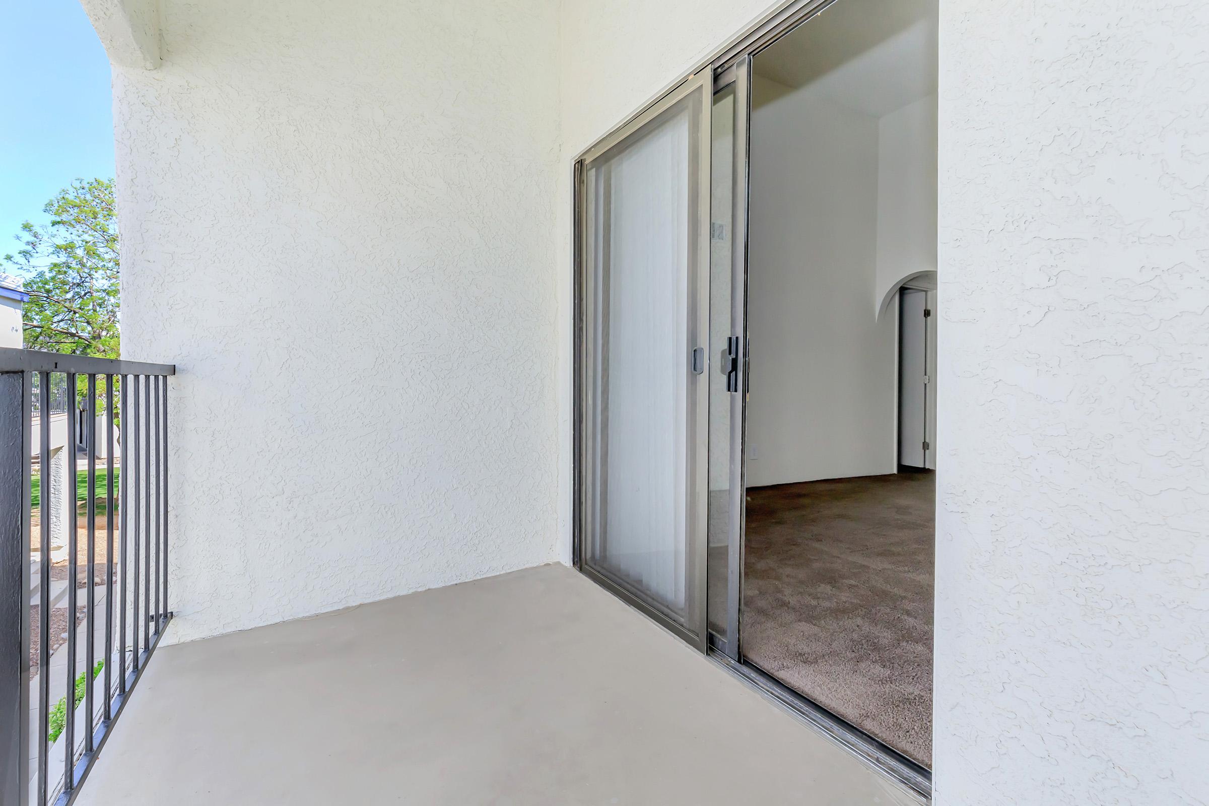 A balcony with a sliding glass door open to an interior room that has carpeted flooring. The wall is painted white, and there are trees visible outside. Natural light is coming in, creating a bright and airy atmosphere.