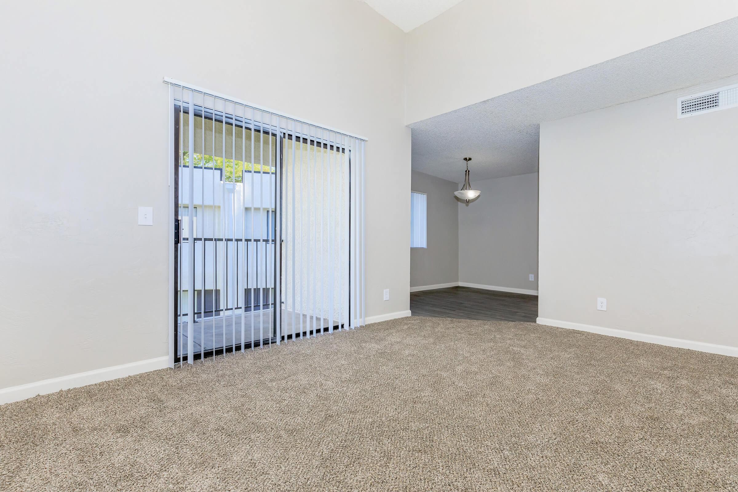 Spacious interior view of a room featuring light-colored walls, plush carpet, and sliding glass doors leading to a balcony. A dining area is visible in the background, illuminated by a pendant light fixture. Natural light streams through nearby windows, creating a bright and inviting atmosphere.