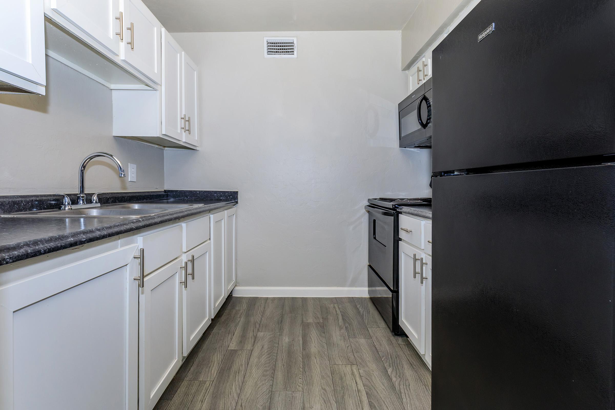 Clean and modern kitchen with white cabinets, a black refrigerator, and a black stove. The countertops are dark, and there is a stainless steel sink. The flooring is a light wood-like laminate, and the walls are painted light gray. Natural light is minimal, and the space is organized with ample storage.
