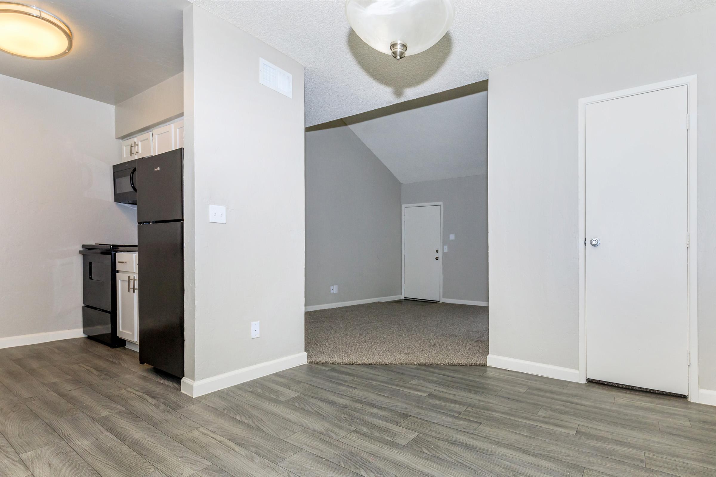 Interior view of an apartment showcasing a kitchen area with black appliances and white cabinets to the left. The space transitions into a carpeted living area with a high ceiling. A door leading to the entrance is visible in the background, and neutral-colored walls enhance the bright, open feel of the space.